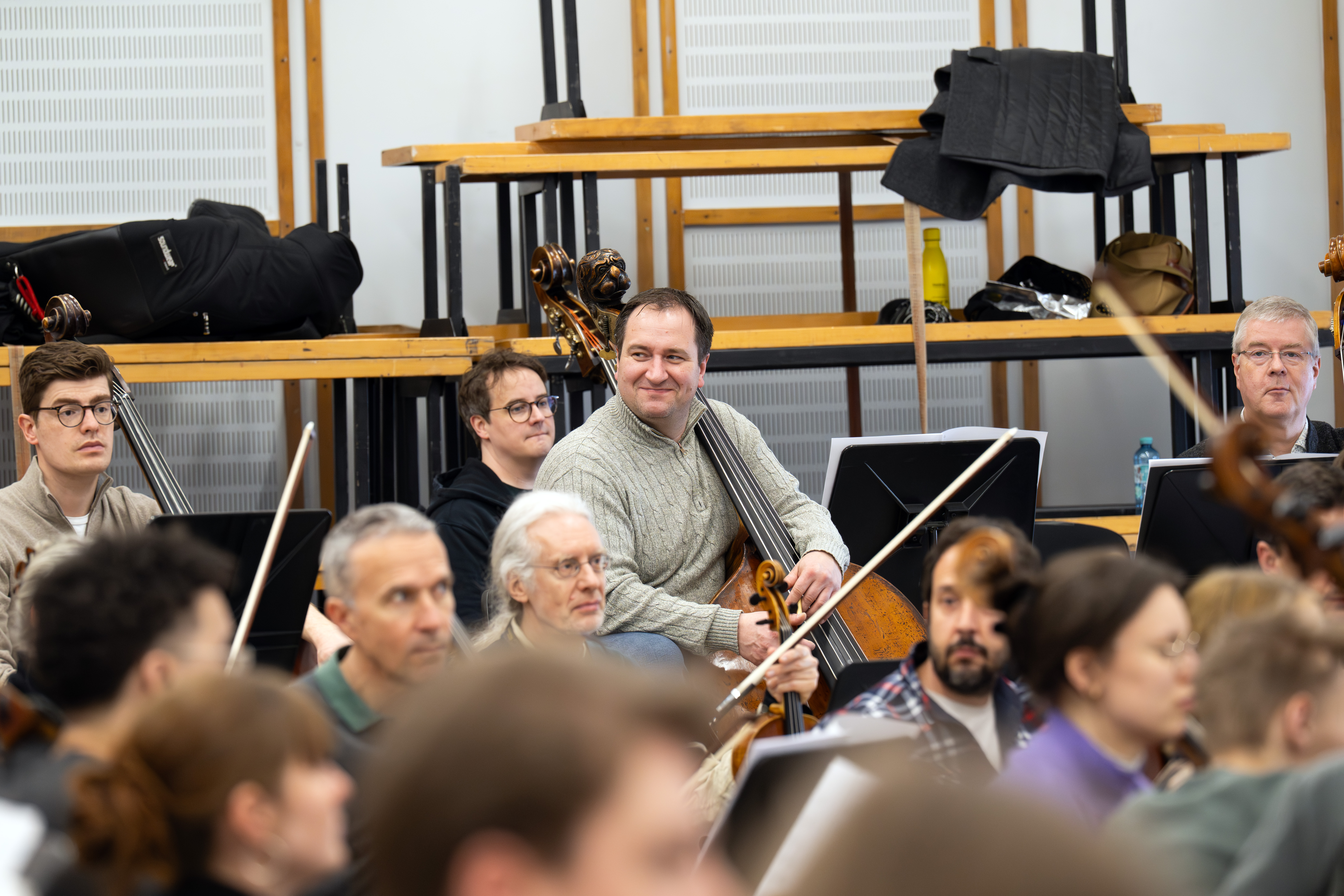 Musiker sitzen mit ihren Instrumenten in einem Proberaum, konzentriert und aufmerksam. Ein Mann in einem grauen Pullover in der Mitte lächelt, während er ein Cello hält. Im Hintergrund sind Regale und Mäntel zu sehen.