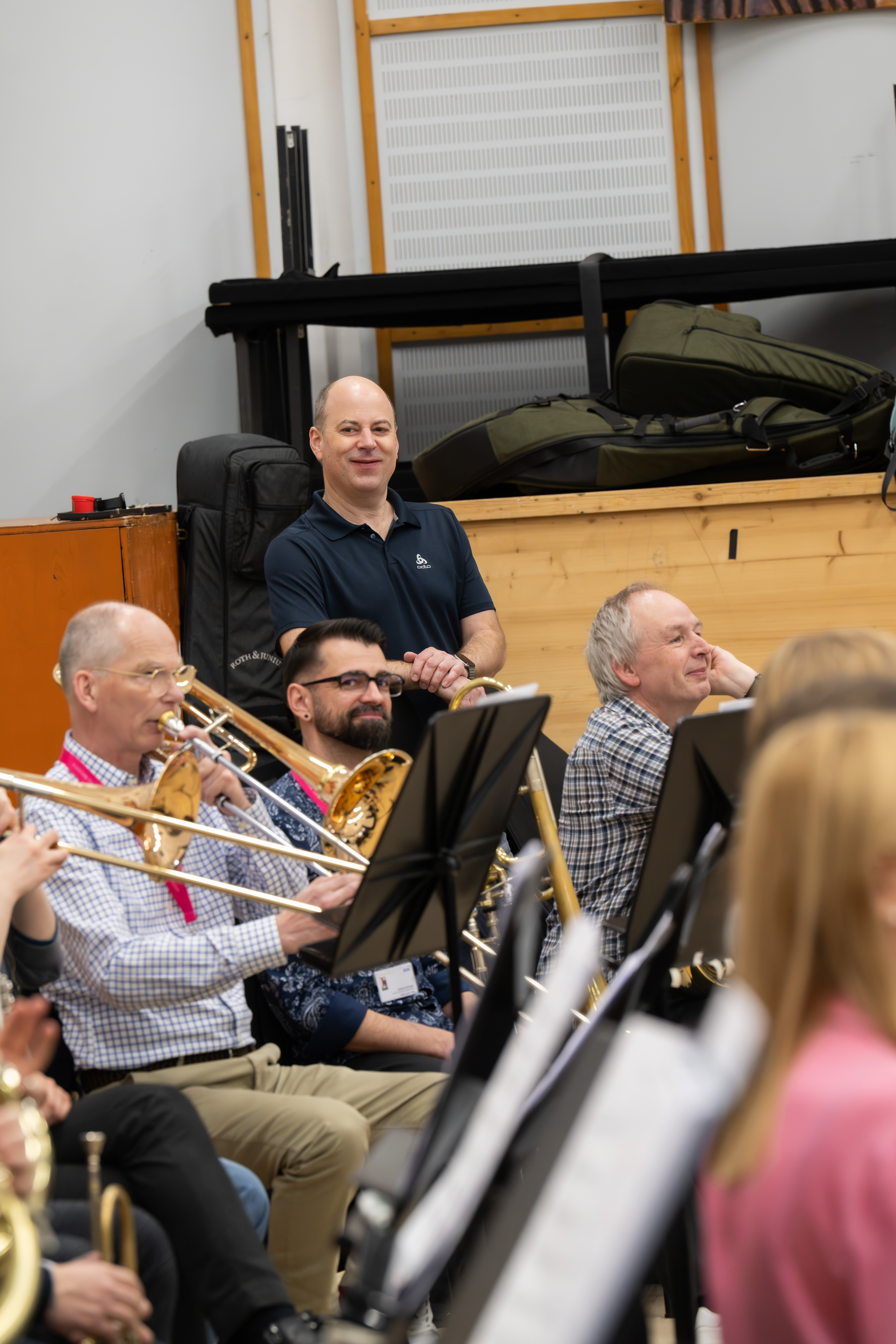 Eine Gruppe von Musikern, spielen Blechblasinstrumente und lächeln während einer Probe in einem Musikraum. Ein Mann, der im Hintergrund steht, schaut zu und lächelt; andere sitzen und scheinen den Moment zu genießen.