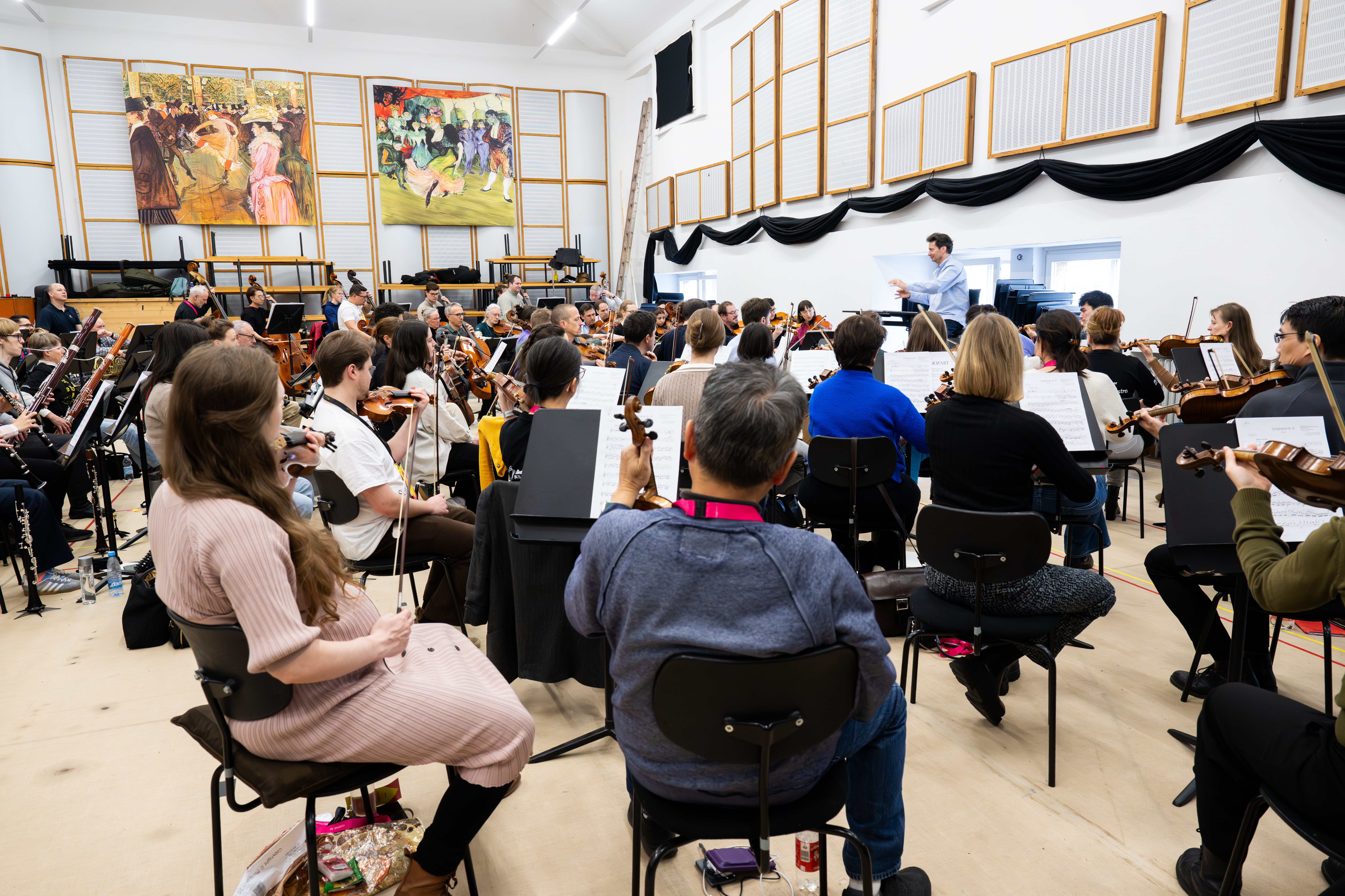 Ein Orchester probt in einem hellen Raum, in dem die Musiker im Halbkreis sitzen und verschiedene Instrumente spielen, während der Dirigent vorne steht. Große Gemälde und Tontafeln schmücken die Wände.