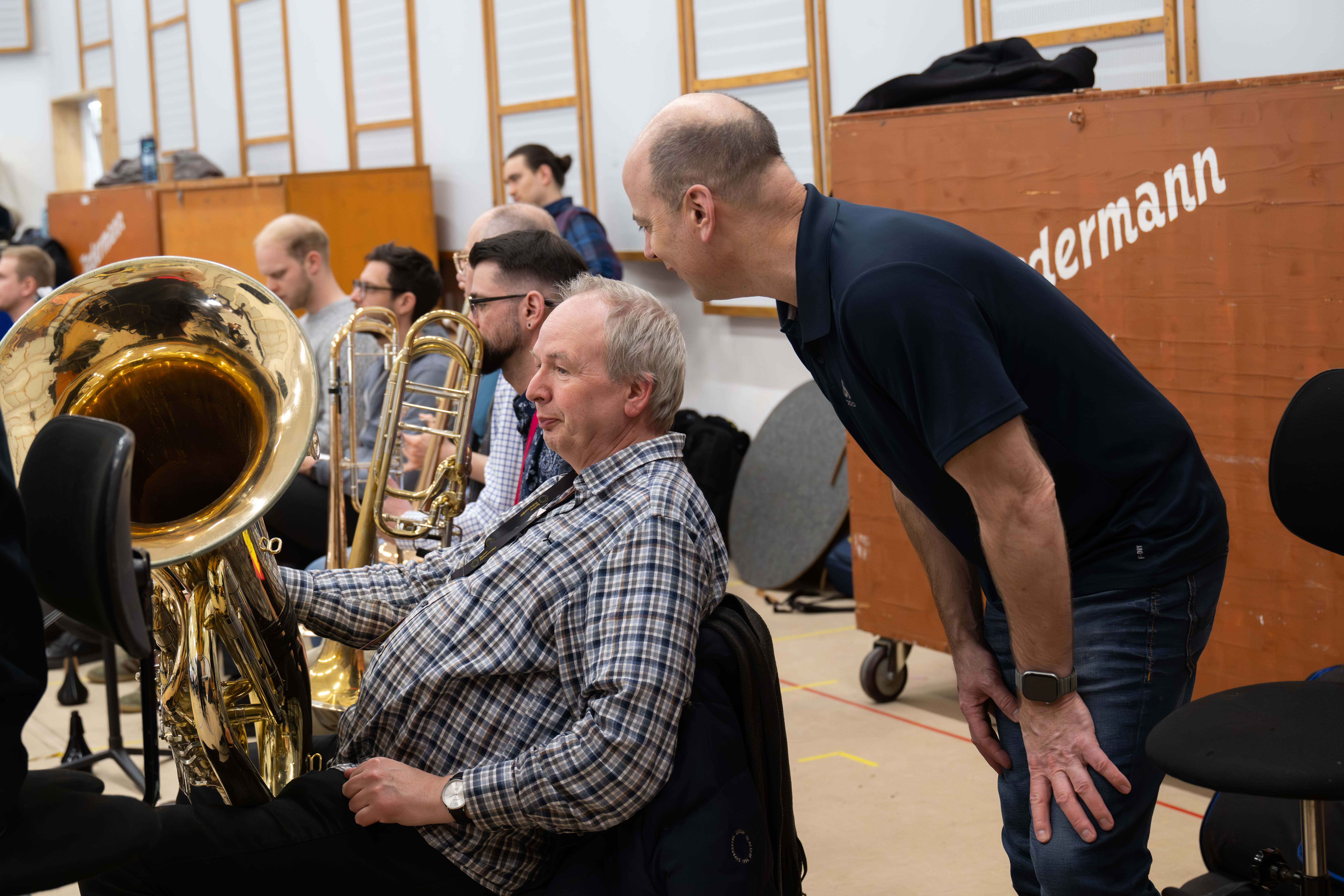 Ein Mann, der ein Blechblasinstrument in der Hand hält, sitzt zwischen anderen Musikern, während sich ein anderer Mann zu ihm hinüberbeugt, um mit ihm in einem Proberaum zu sprechen. Im Hintergrund sind Notenständer und Holztafeln zu sehen.