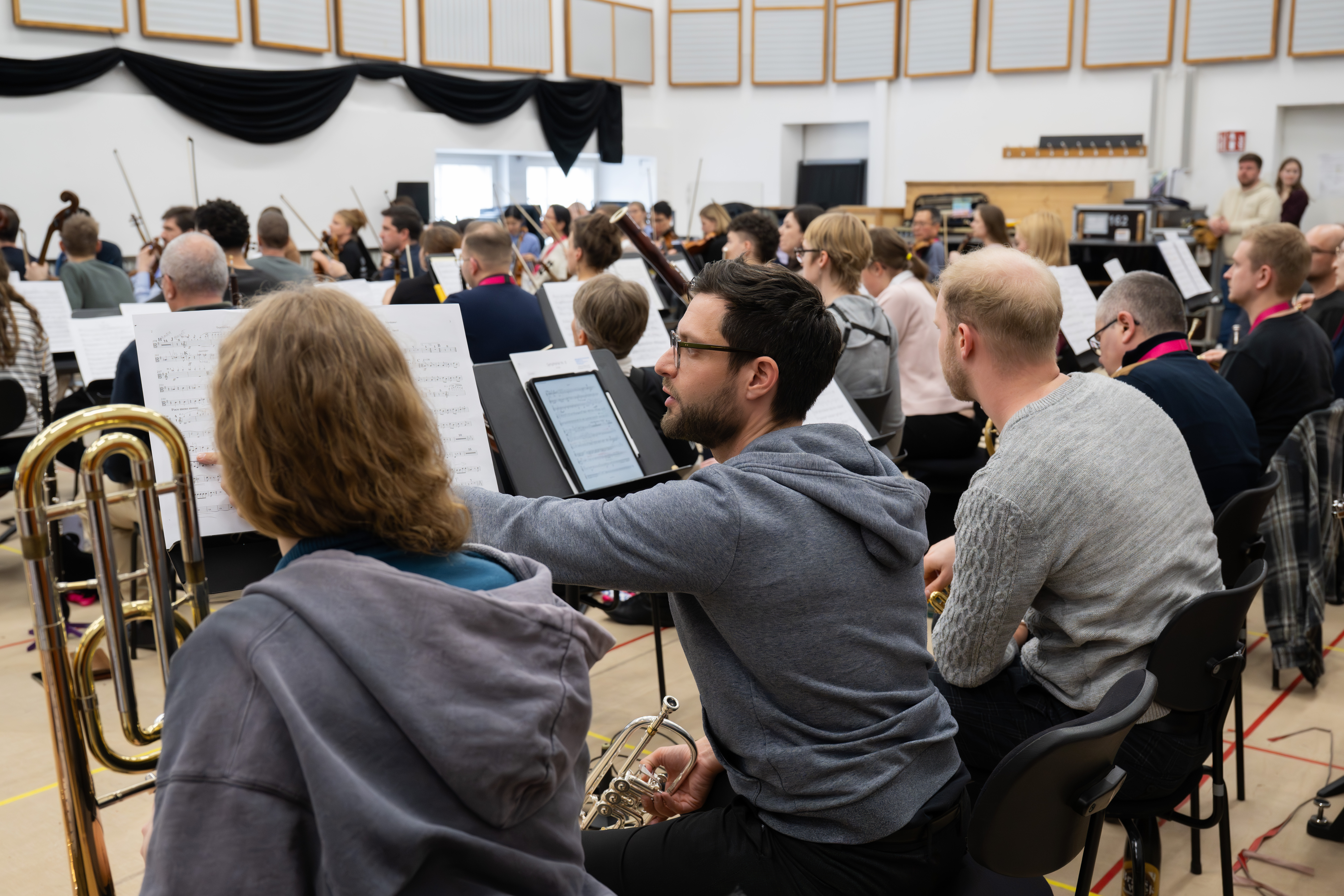 Eine Gruppe von Musikern probt in einem hellen Raum. Sie sitzen in Reihen mit Notenständern und Instrumenten. Eine Person zeigt auf die Notenblätter und unterhält sich mit ihrem Sitznachbarn.
