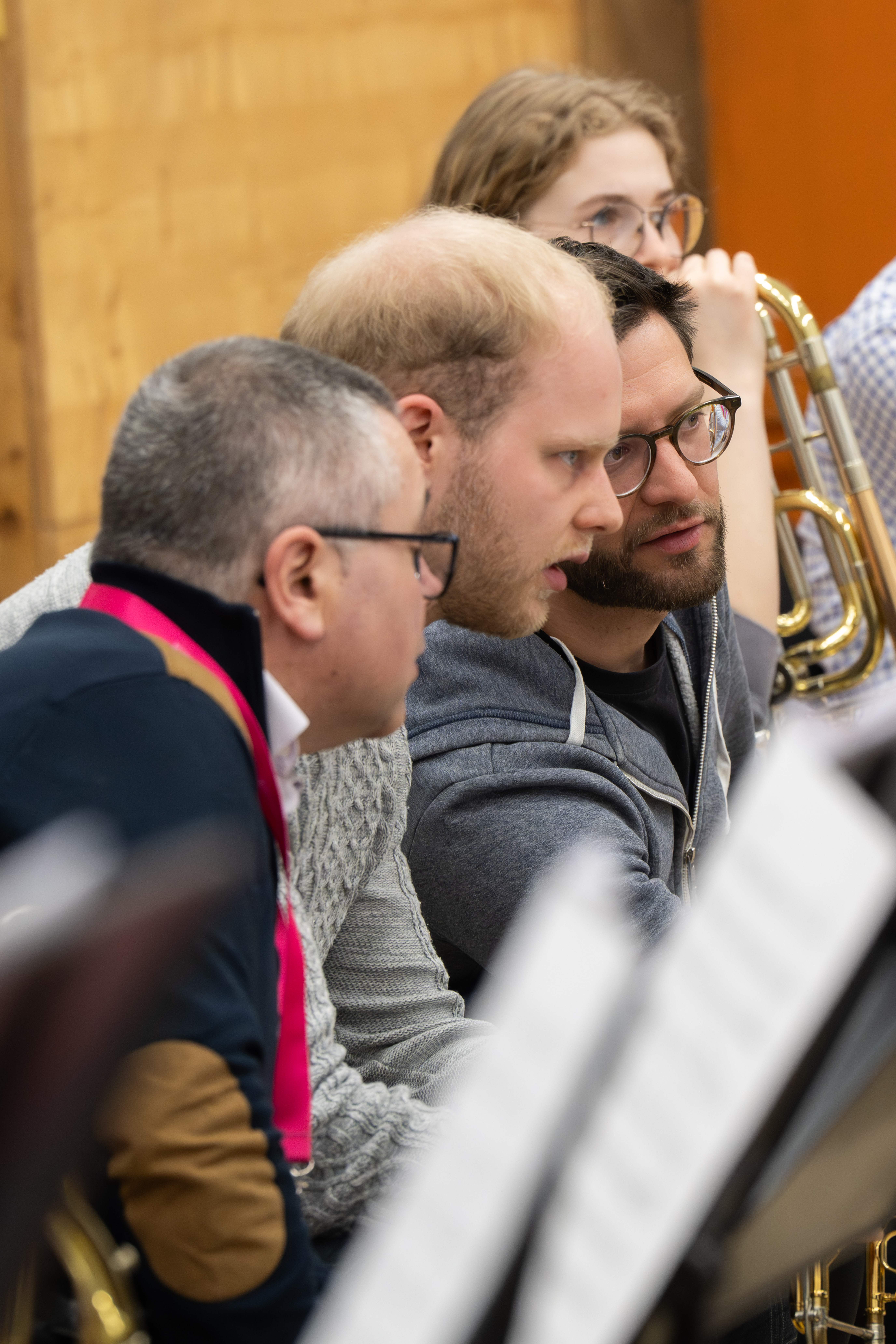Drei Männer sitzen eng beieinander und unterhalten sich angeregt, vor ihnen stehen Notenständer. Im Hintergrund ist eine Frau zu sehen, die ein Blechblasinstrument hält. Die Umgebung scheint ein Proberaum zu sein.