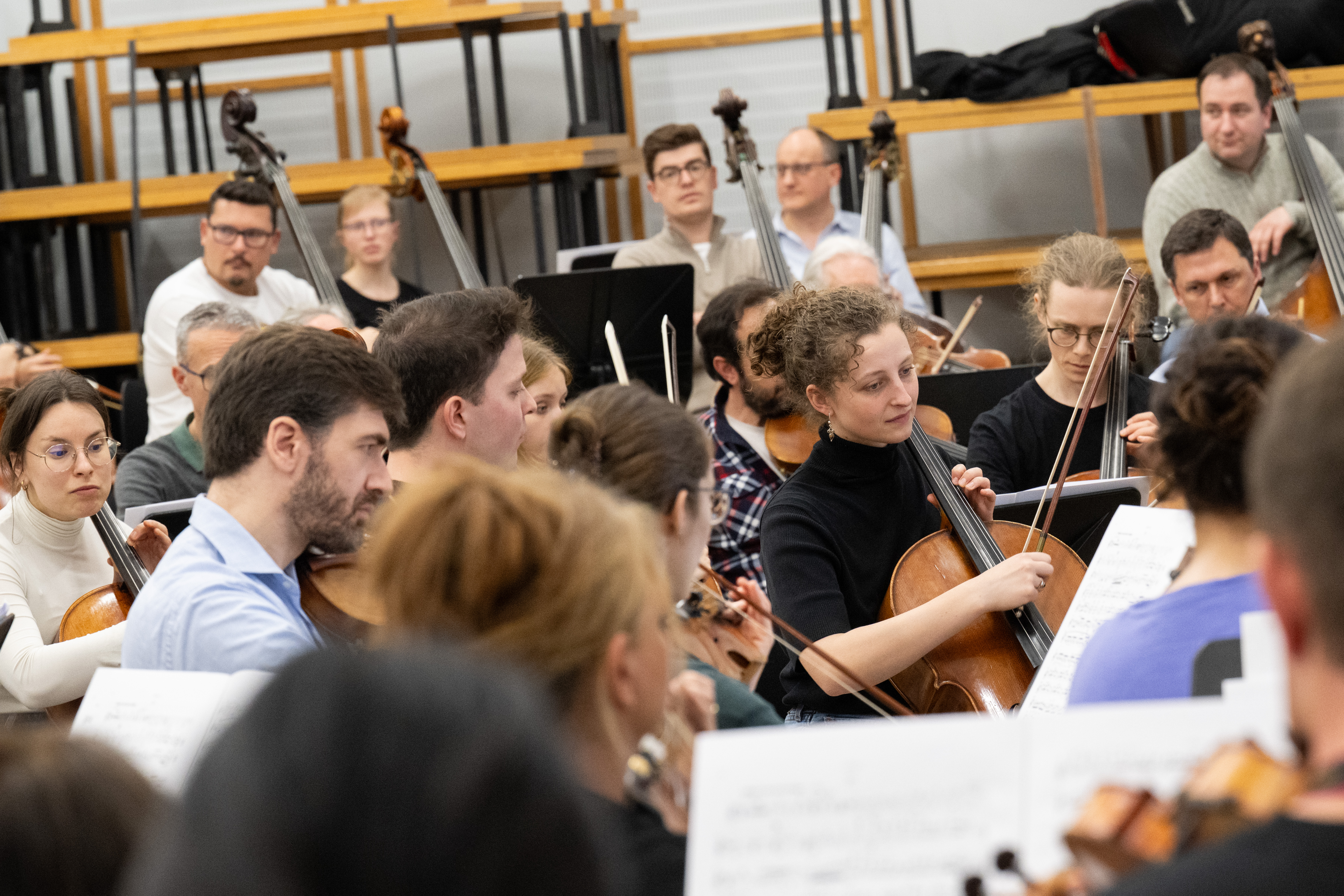 Eine Gruppe von Musikern probt in einer Orchesterumgebung, spielt Streichinstrumente und liest Noten, während im Hintergrund leere Sitzreihen zu sehen sind.
