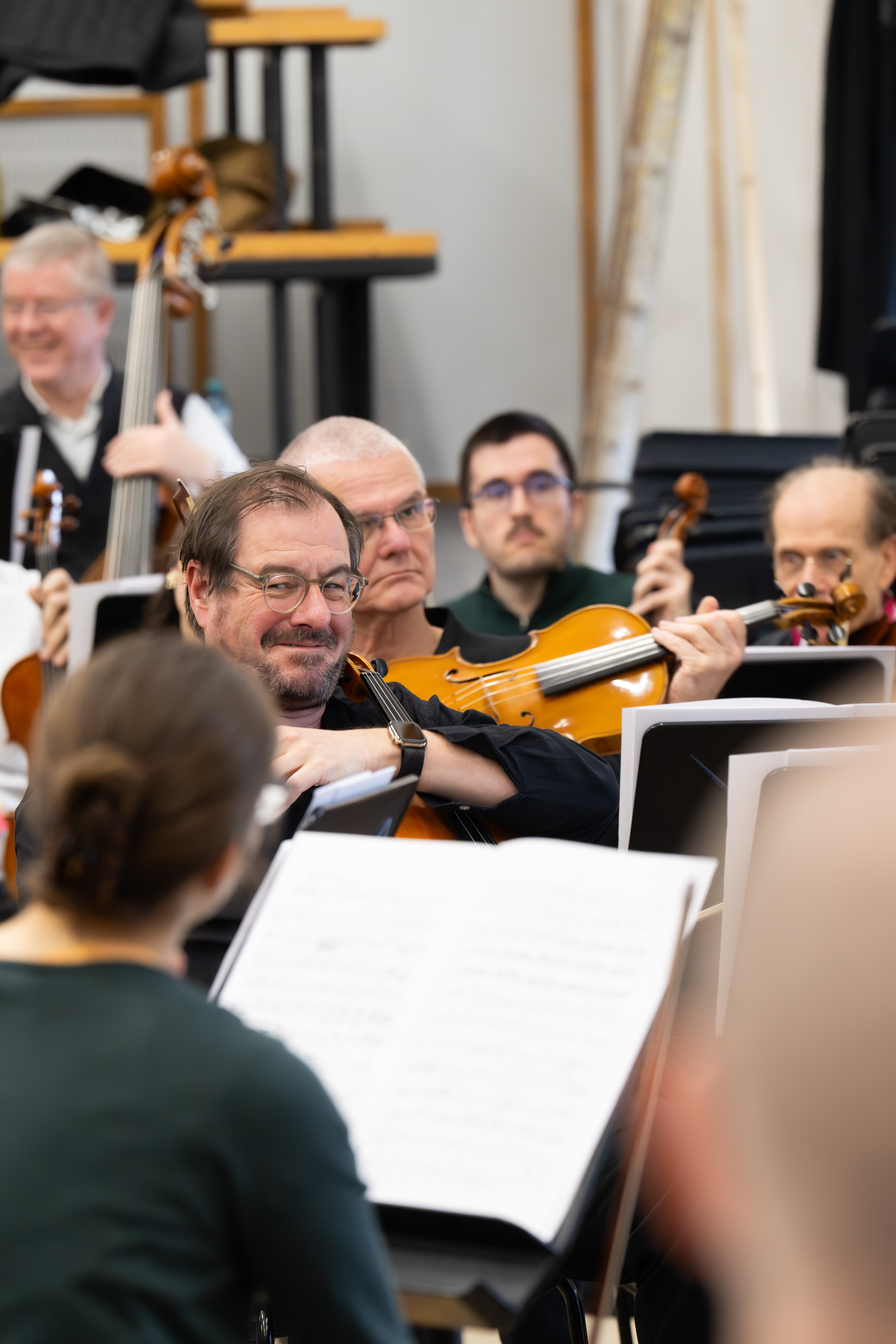 Eine Gruppe von Musikern bei einer Orchesterprobe, die sich auf einen lächelnden Mann konzentriert, der Bratsche spielt. Im Vordergrund sind Notenständer mit Notenblättern zu sehen.