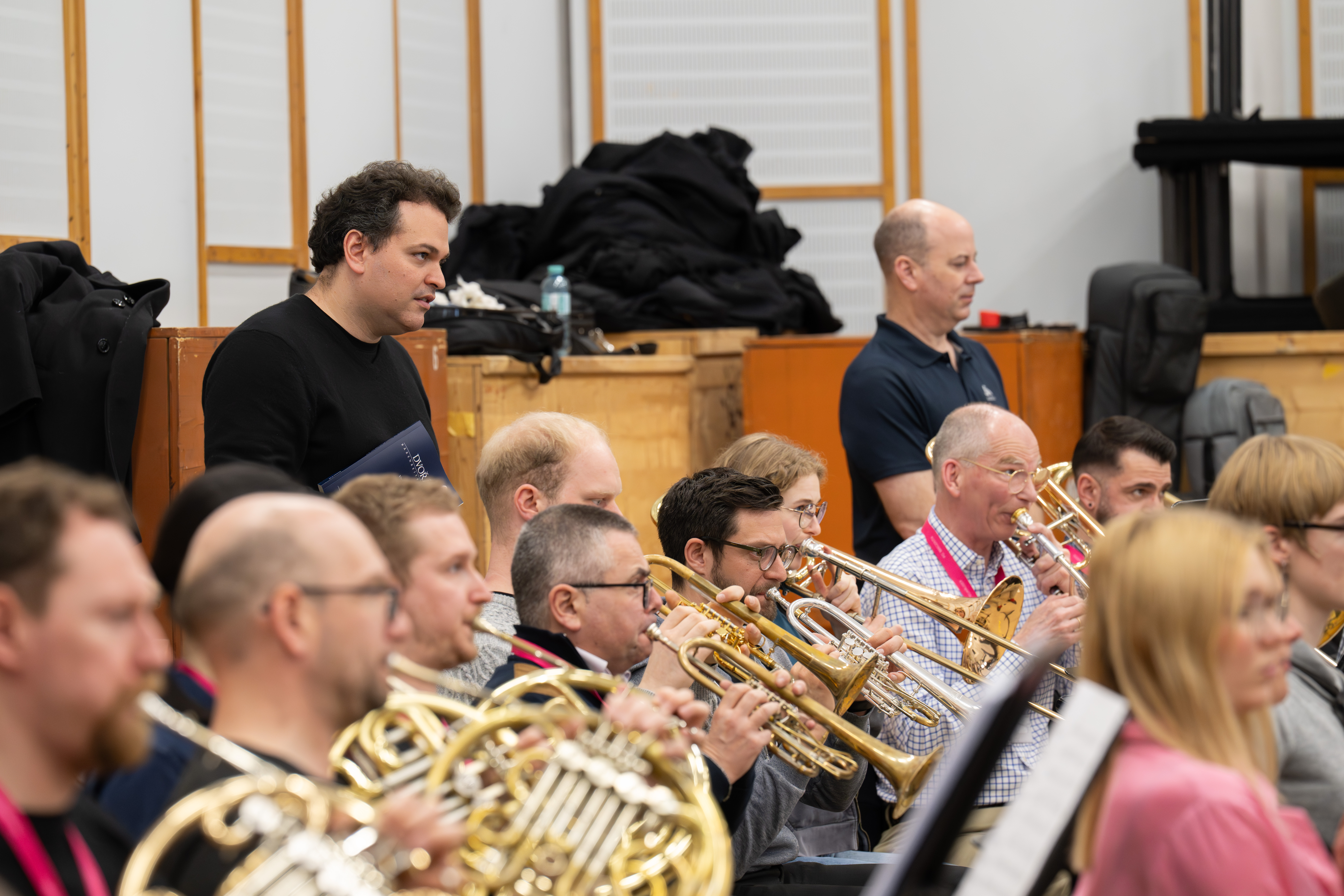 Eine Gruppe von Musikern spielt Blechblasinstrumente in einem Proberaum. Zwei Männer stehen hinter ihnen und beobachten, während die sitzenden Musiker sich auf ihre Notenblätter und Instrumente konzentrieren.