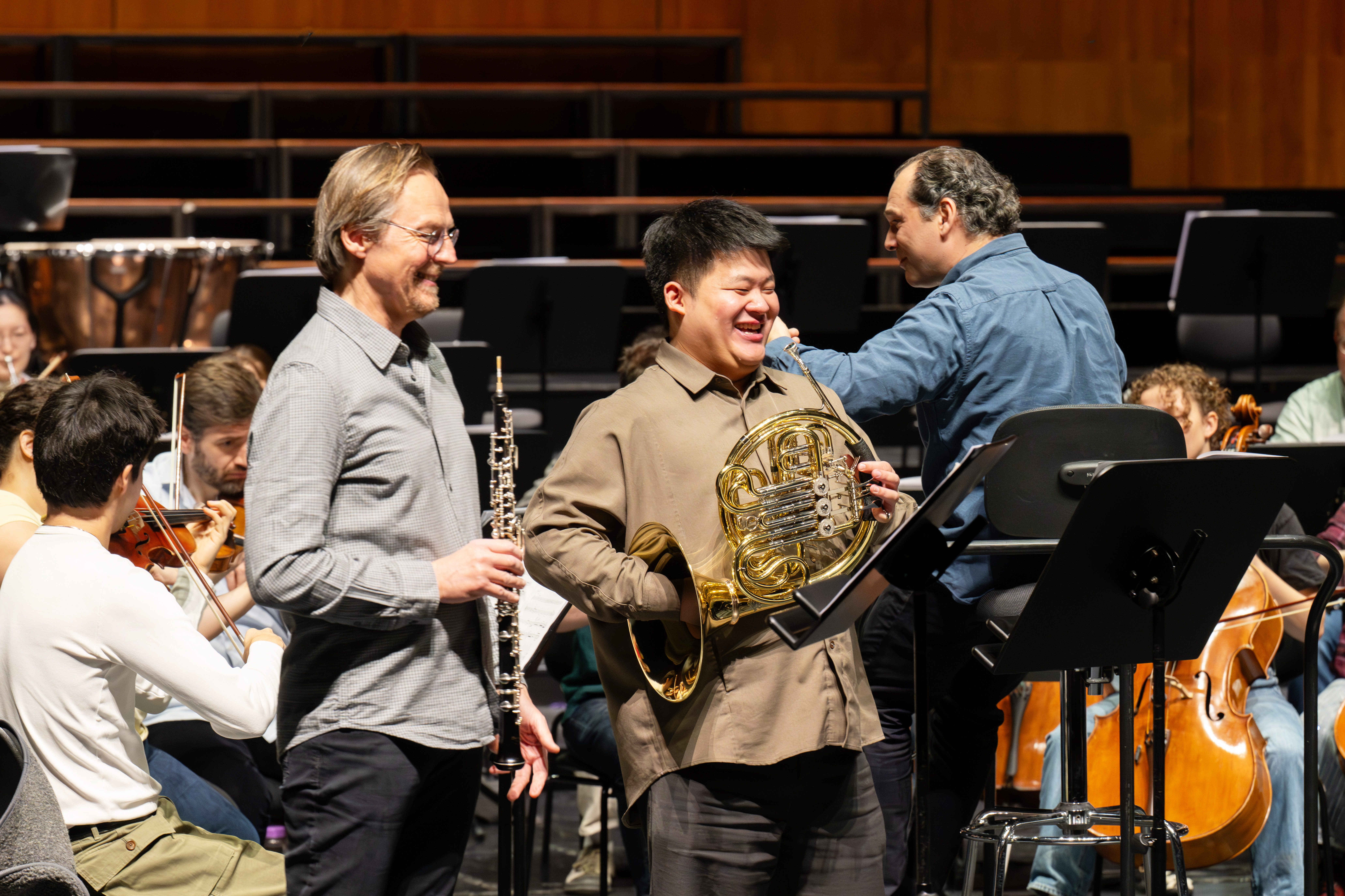Eine Gruppe von Musikern probt auf der Bühne. Zwei lächelnde Männer im Vordergrund halten eine Oboe und ein Waldhorn. Andere Musiker spielen im Hintergrund Streichinstrumente. Notenständer sind zu sehen.