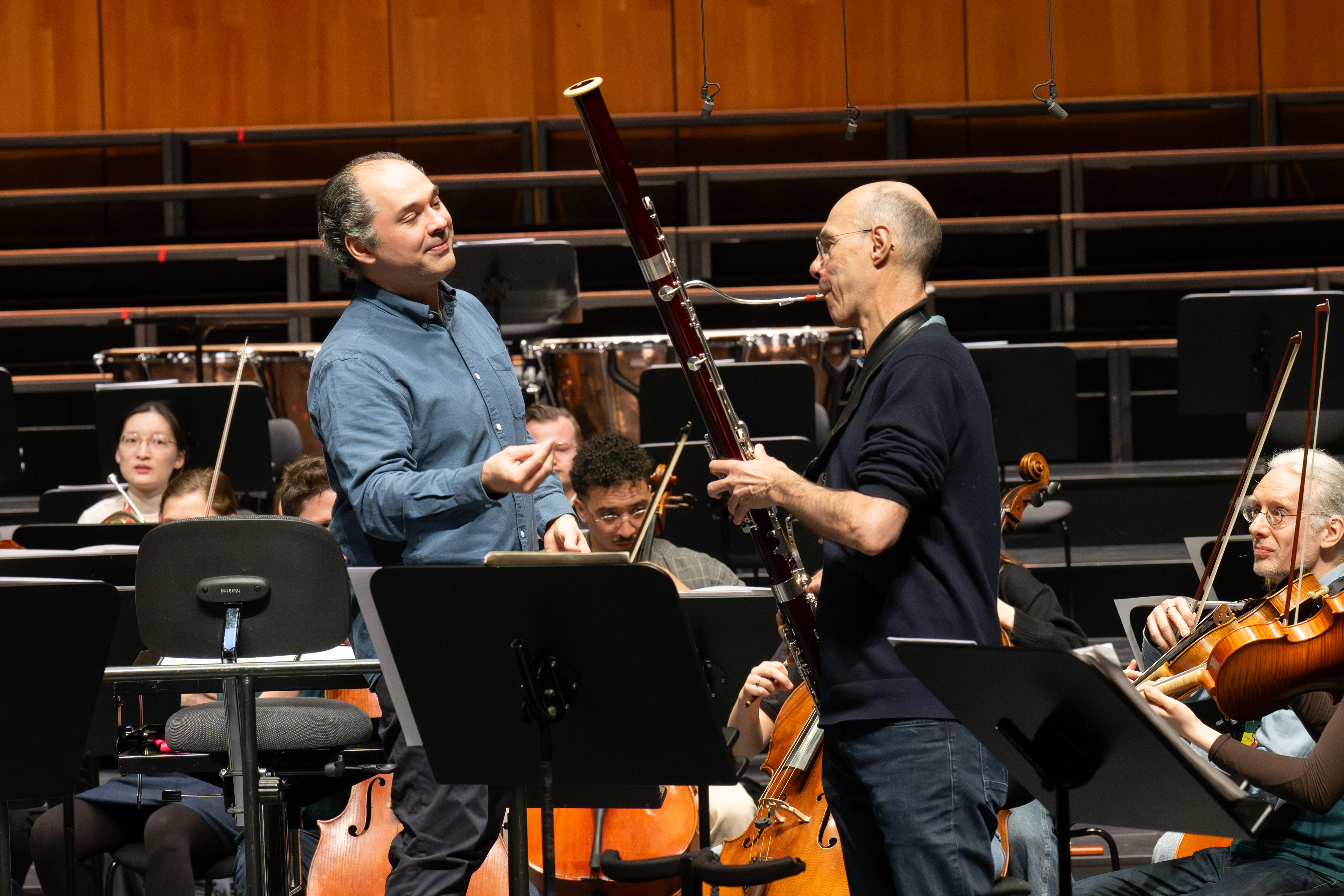 Ein Dirigent im blauen Hemd gestikuliert, während er während einer Orchesterprobe mit einem Fagottisten spricht, während im Hintergrund andere Musiker und Streichinstrumente zu sehen sind.