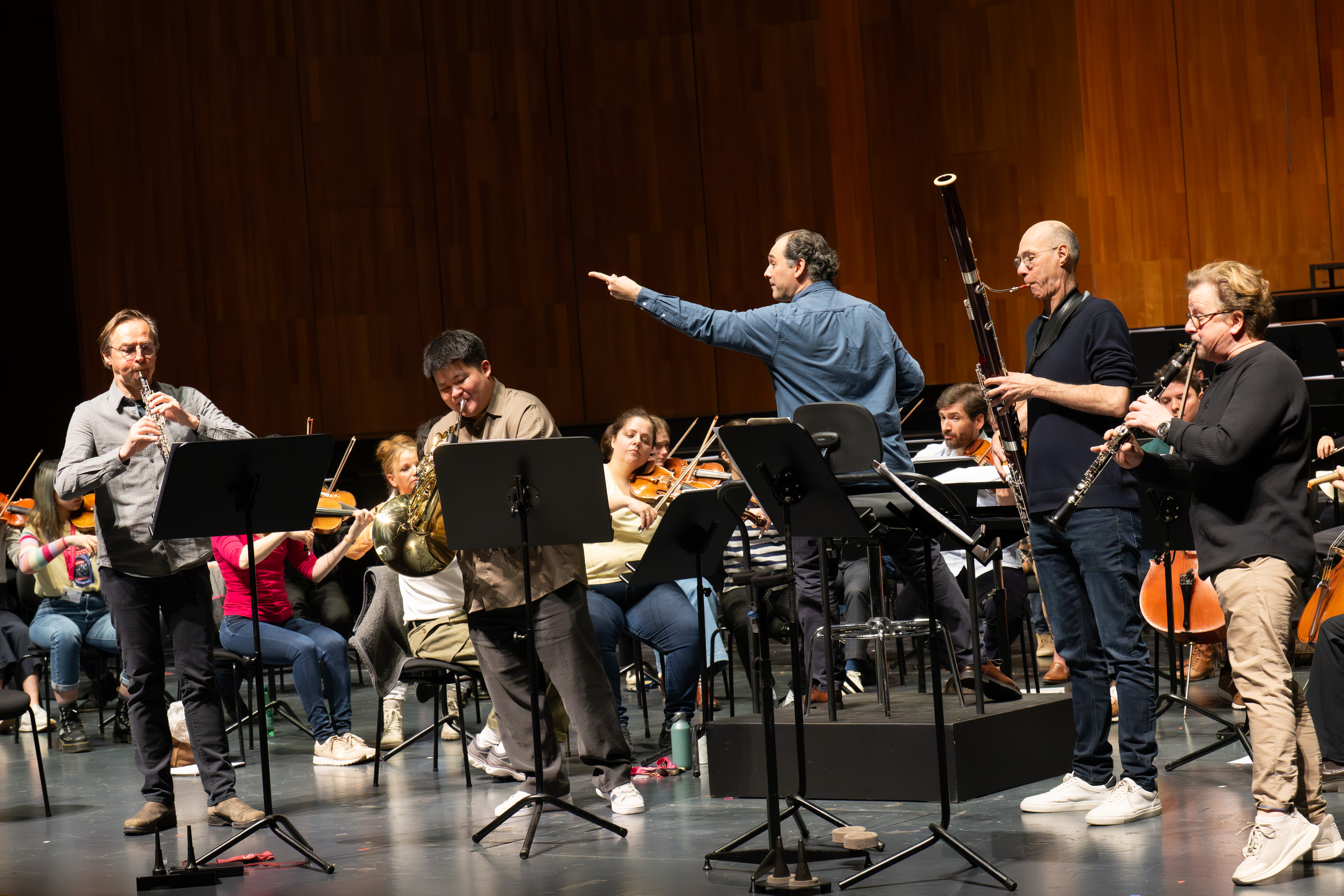 Ein Dirigent leitet eine Orchesterprobe auf der Bühne, bei der vier Holz- und Blechbläsersolisten vor sitzenden Musikern und Notenständern vor einem hölzernen Hintergrund stehen und spielen.