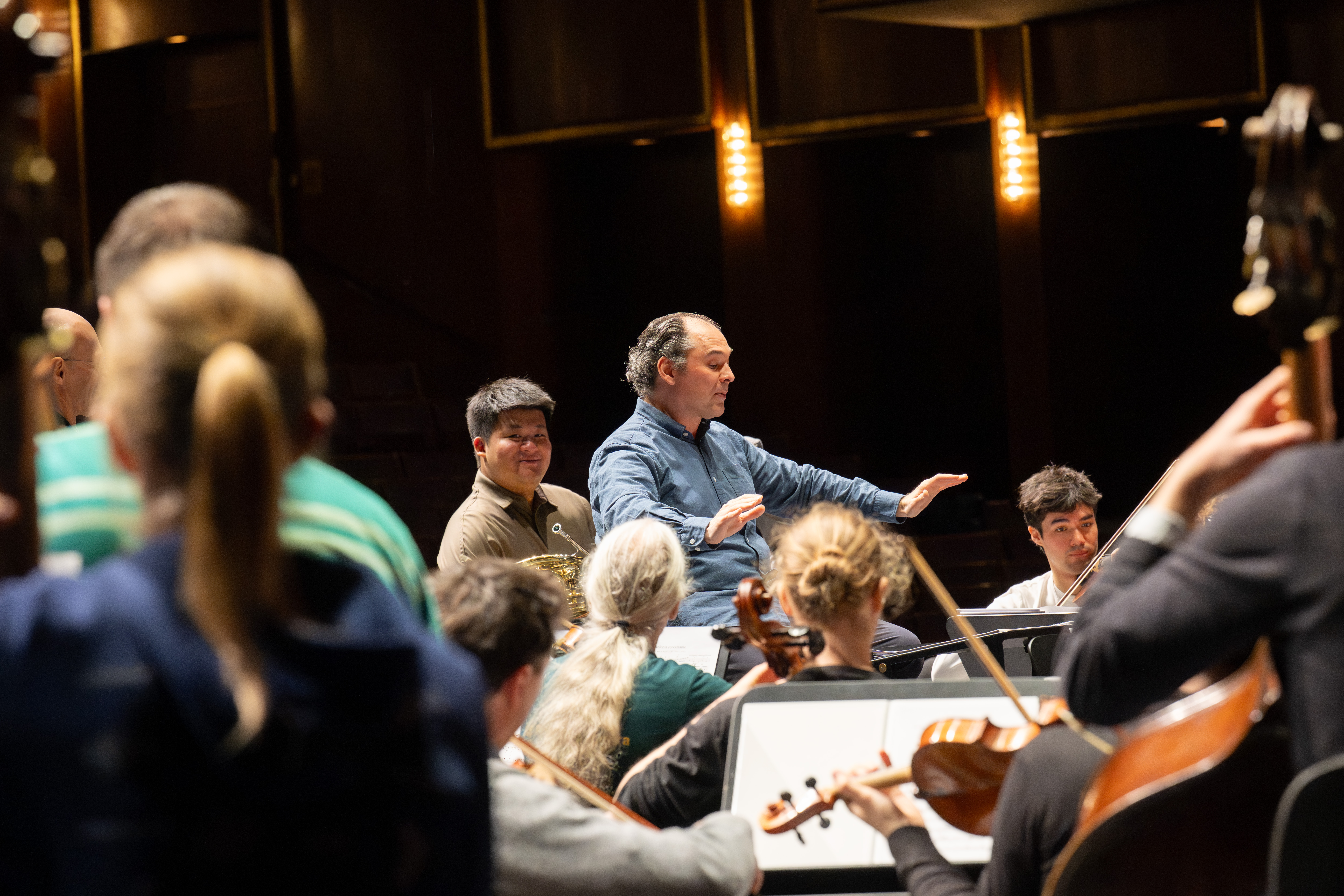 Ein Dirigent leitet ein Orchester während einer Probe. Die Musiker spielen verschiedene Streich- und Blechblasinstrumente, und die Szene wird durch warmes Licht beleuchtet.