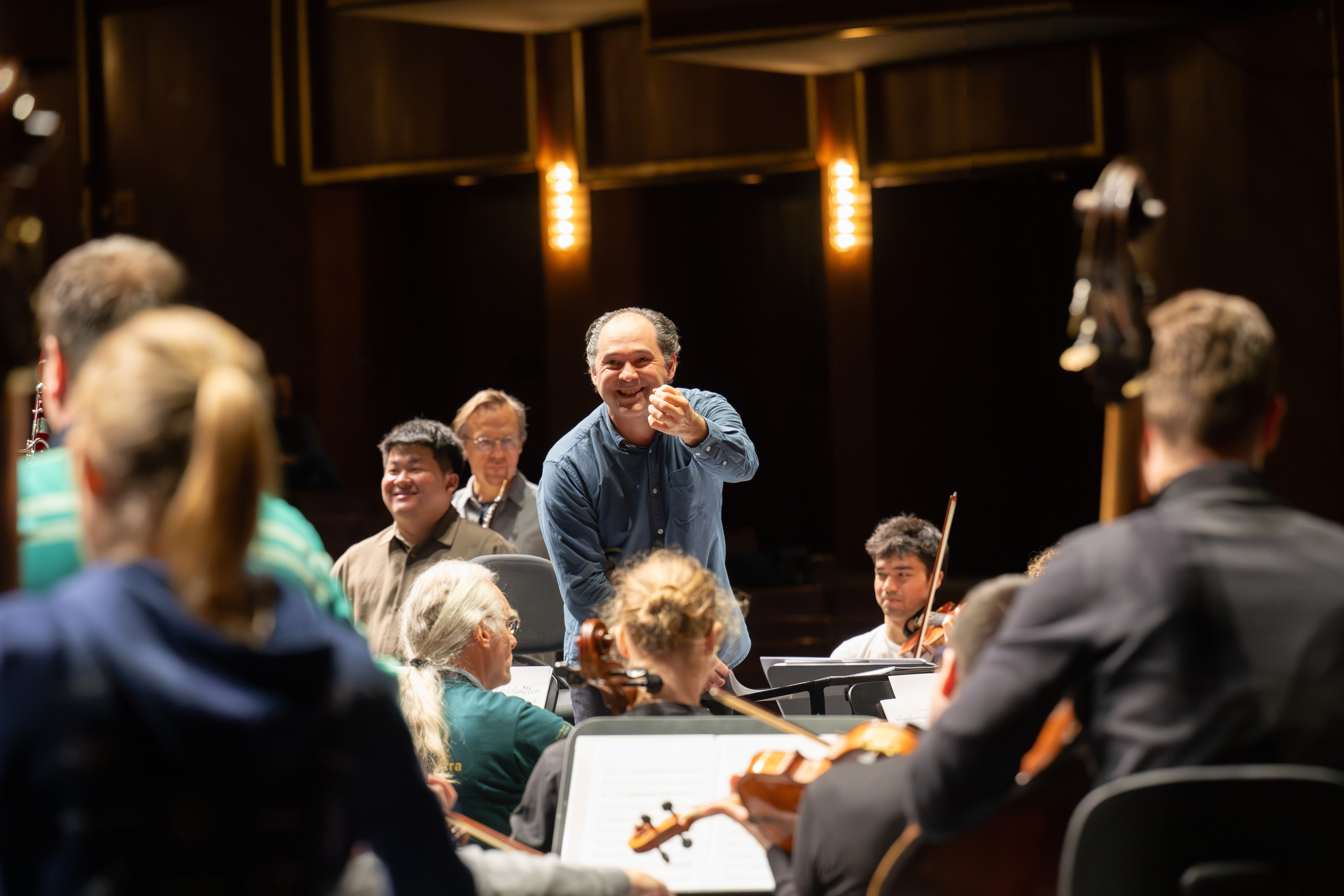 Ein Dirigent steht lächelnd und gestikulierend in Richtung eines Orchesters während einer Probe, die Musiker sitzen, halten ihre Instrumente und warmes Licht beleuchtet die Szene.