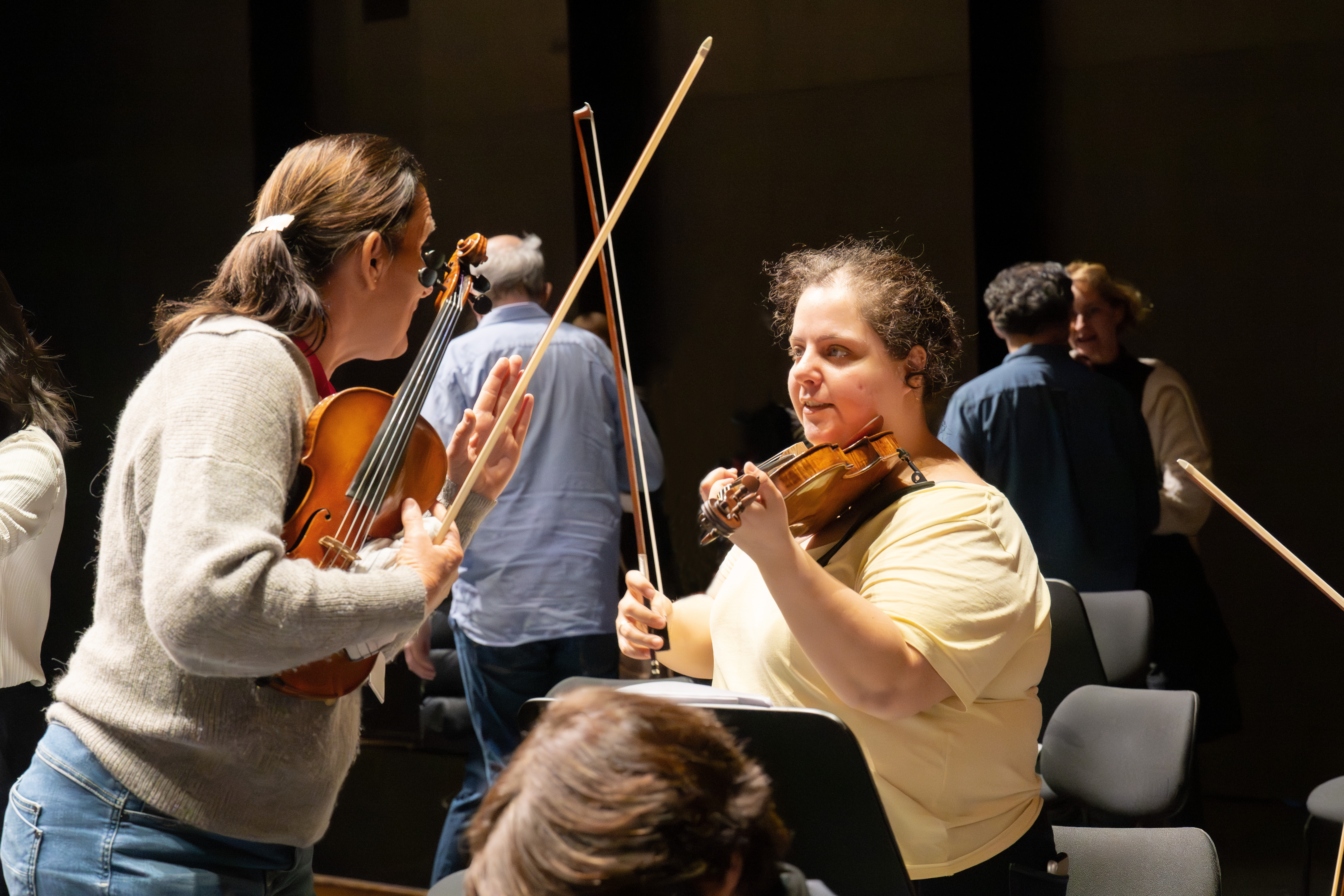 Zwei Frauen mit Geigen und Bögen stehen sich gegenüber und scheinen sich zu unterhalten oder zu proben. Andere Personen stehen im Hintergrund in einem schwach beleuchteten Raum mit leeren Stühlen.