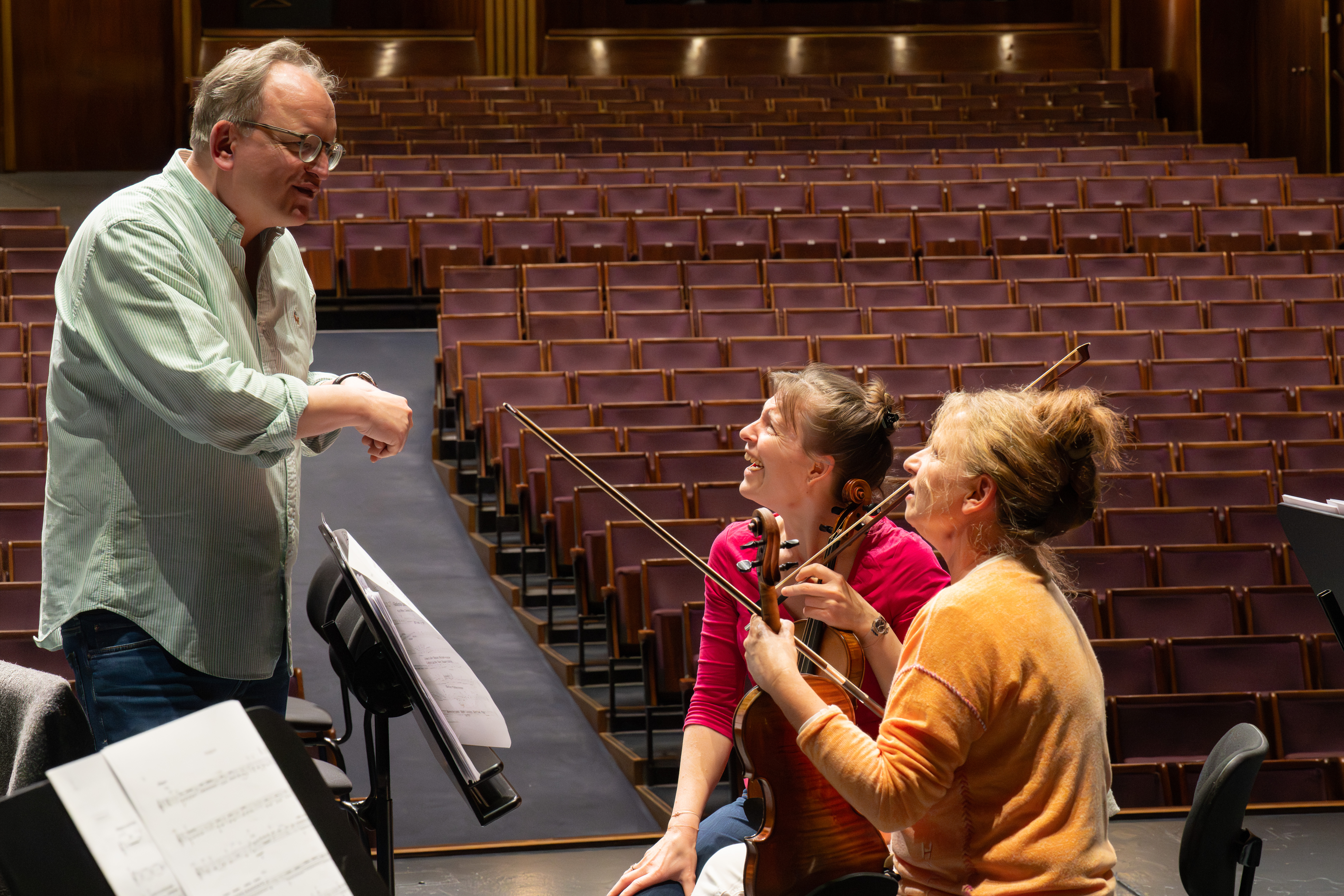 Ein Mann spricht zu zwei lächelnden Geigerinnen, die ihre Instrumente auf einer Bühne in einem leeren Theater mit Reihen brauner Sitze hinter ihnen halten. Im Vordergrund sind Notenblätter zu sehen.
