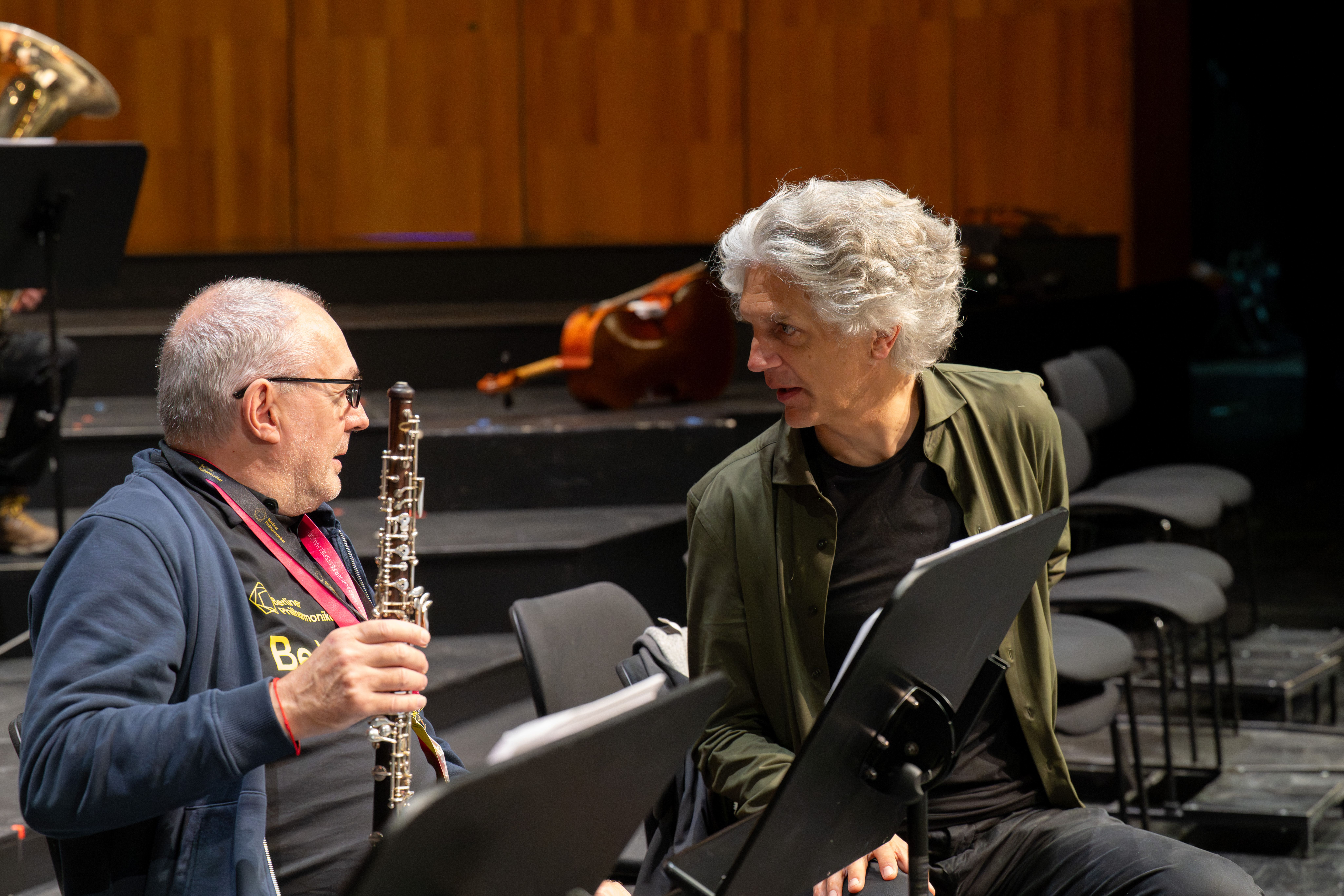Zwei Männer sitzen zwischen Notenständern und Stühlen auf einer Bühne. Einer hält eine Oboe in der Hand und spricht mit dem anderen, der aufmerksam zuhört. Im Hintergrund sind ein Kontrabass und eine Tuba zu sehen.