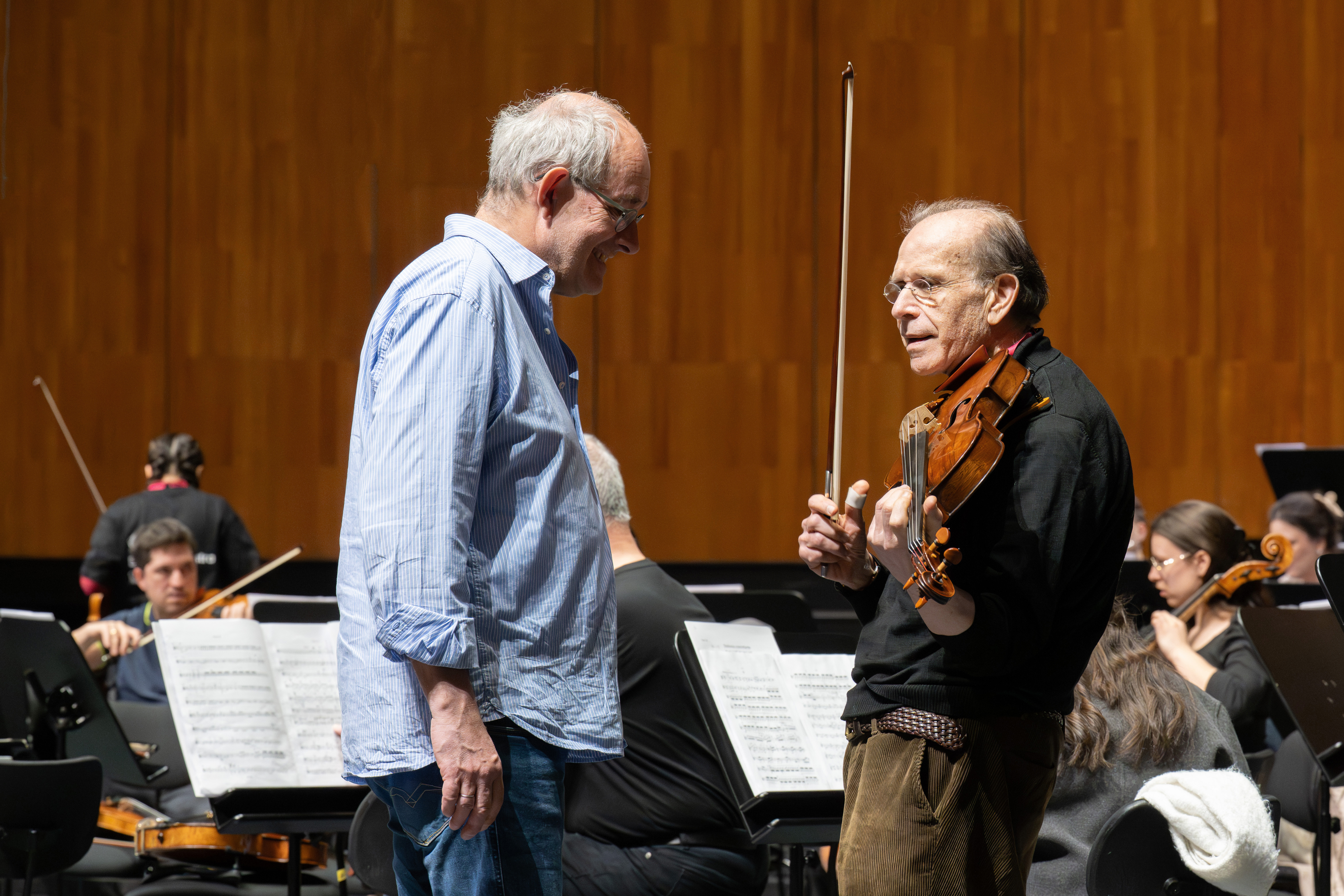 Zwei Männer stehen auf der Bühne und unterhalten sich, einer hält eine Bratsche und einen Bogen. Hinter ihnen sitzen Musiker mit Notenständern und Instrumenten und proben vor einer Holzwand.