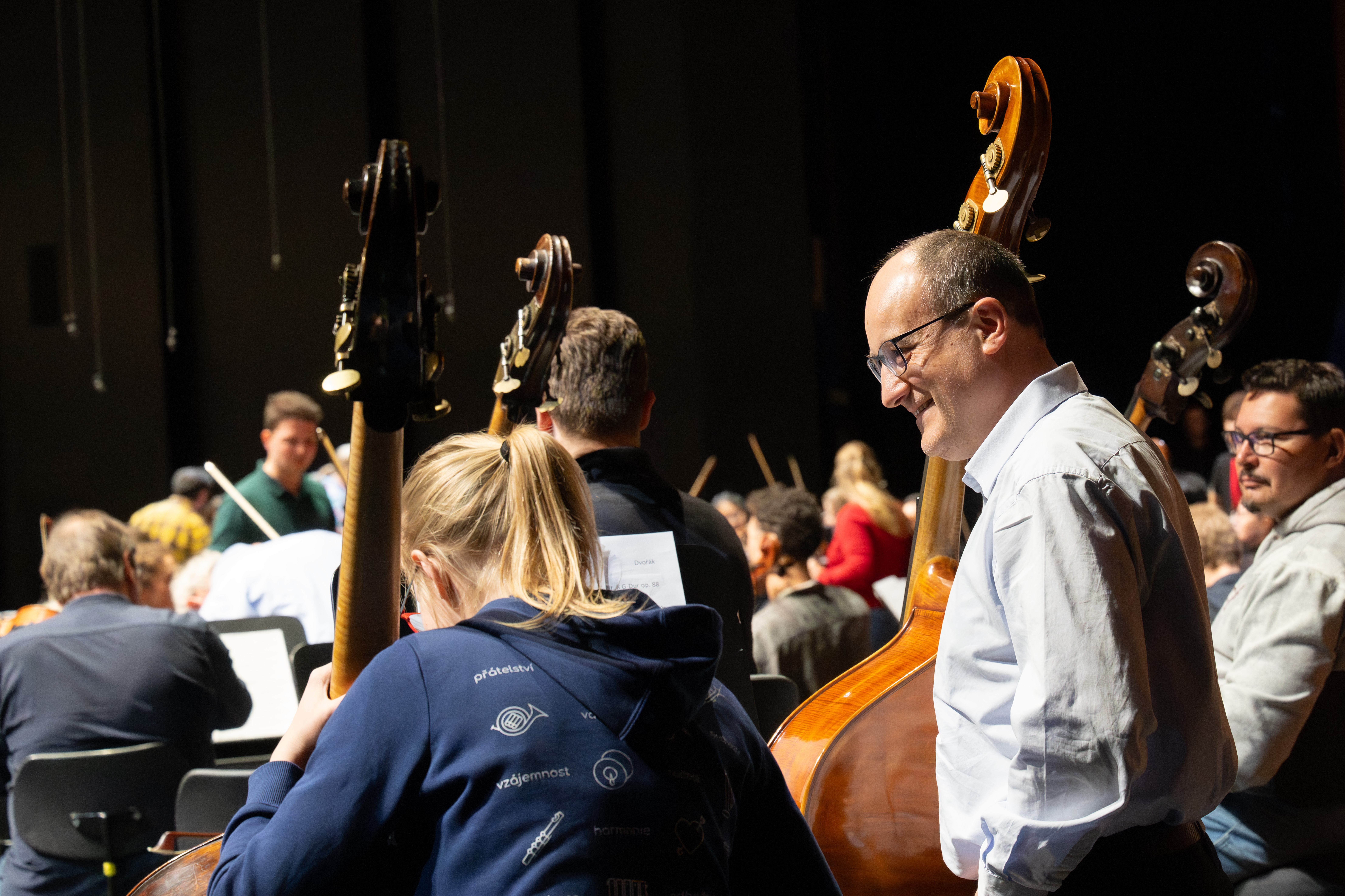Musiker bei einer Orchesterprobe, wobei ein lächelnder Mann mit einem Kontrabass im Mittelpunkt steht. Um ihn herum sind andere Musiker und Instrumente im hellen Bühnenlicht zu sehen.