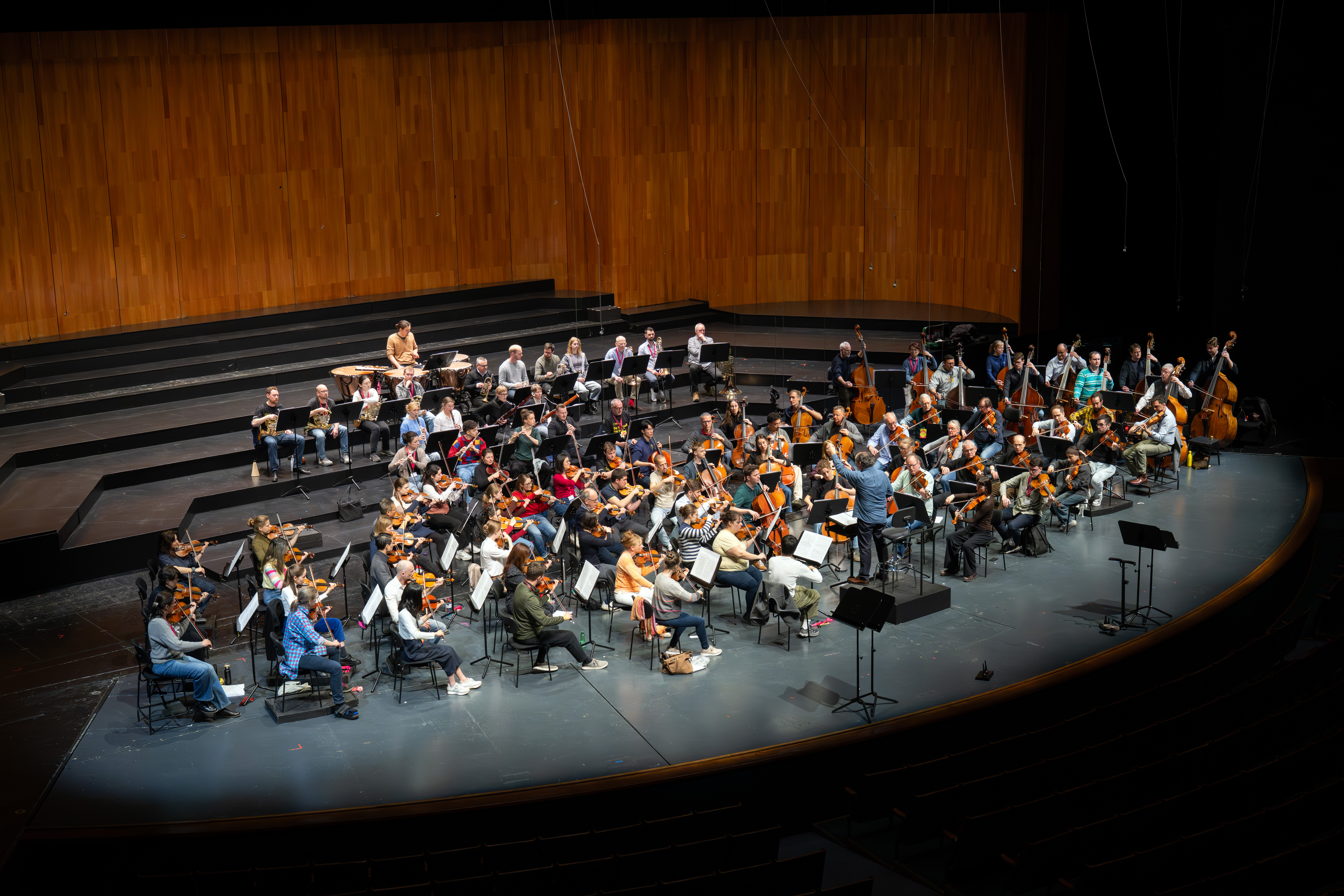 Ein großes Orchester probt auf einer breiten, geschwungenen Bühne mit abgestuften Sitzreihen dahinter. Die Musiker spielen Streich-, Blech- und Holzblasinstrumente, während ein Dirigent von vorne dirigiert.