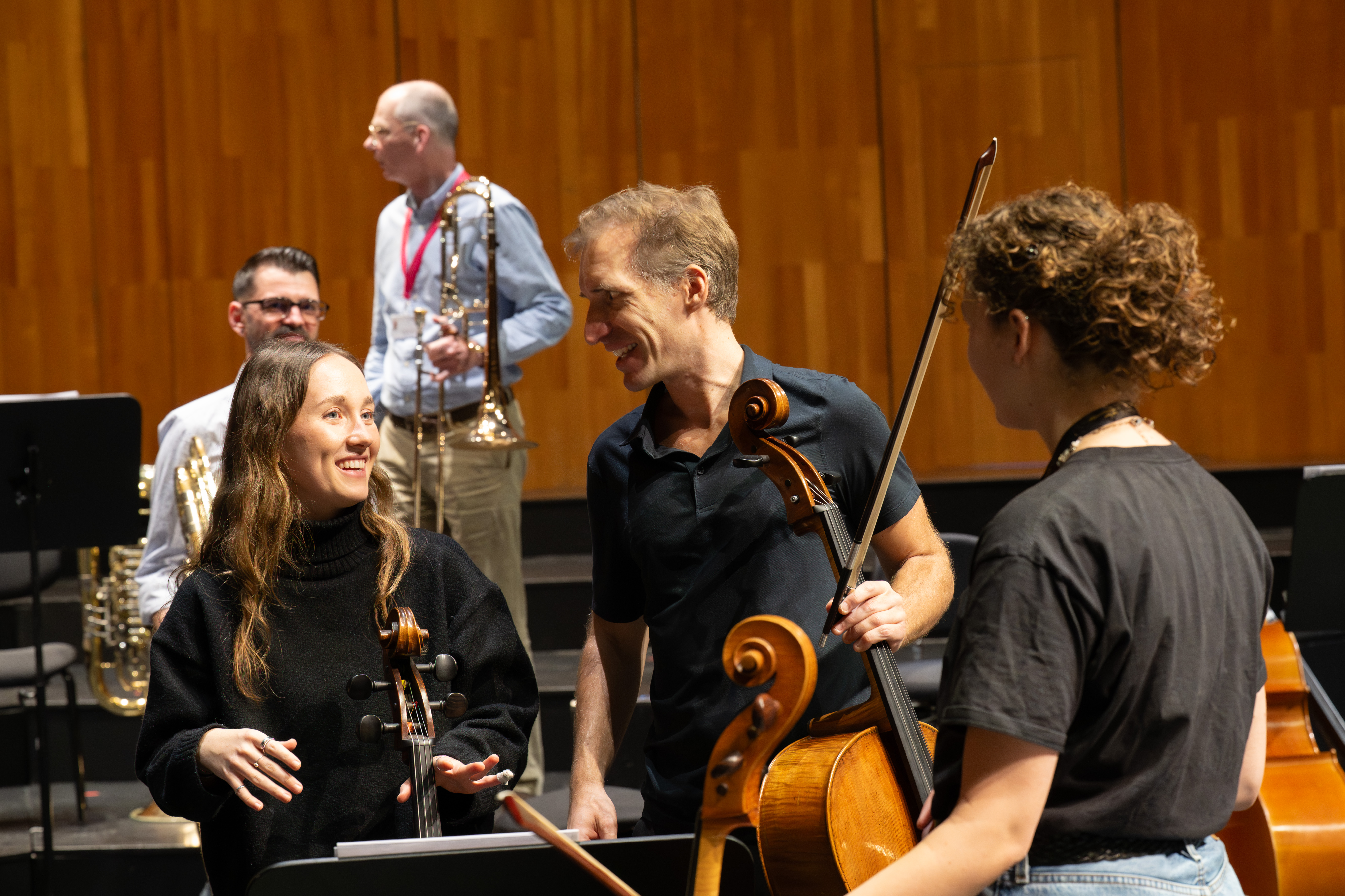 Drei Musiker mit Streichinstrumenten unterhalten sich auf der Bühne und lächeln miteinander, während zwei Blechbläser im Hintergrund an einer Holzwand in einer Probenumgebung stehen.