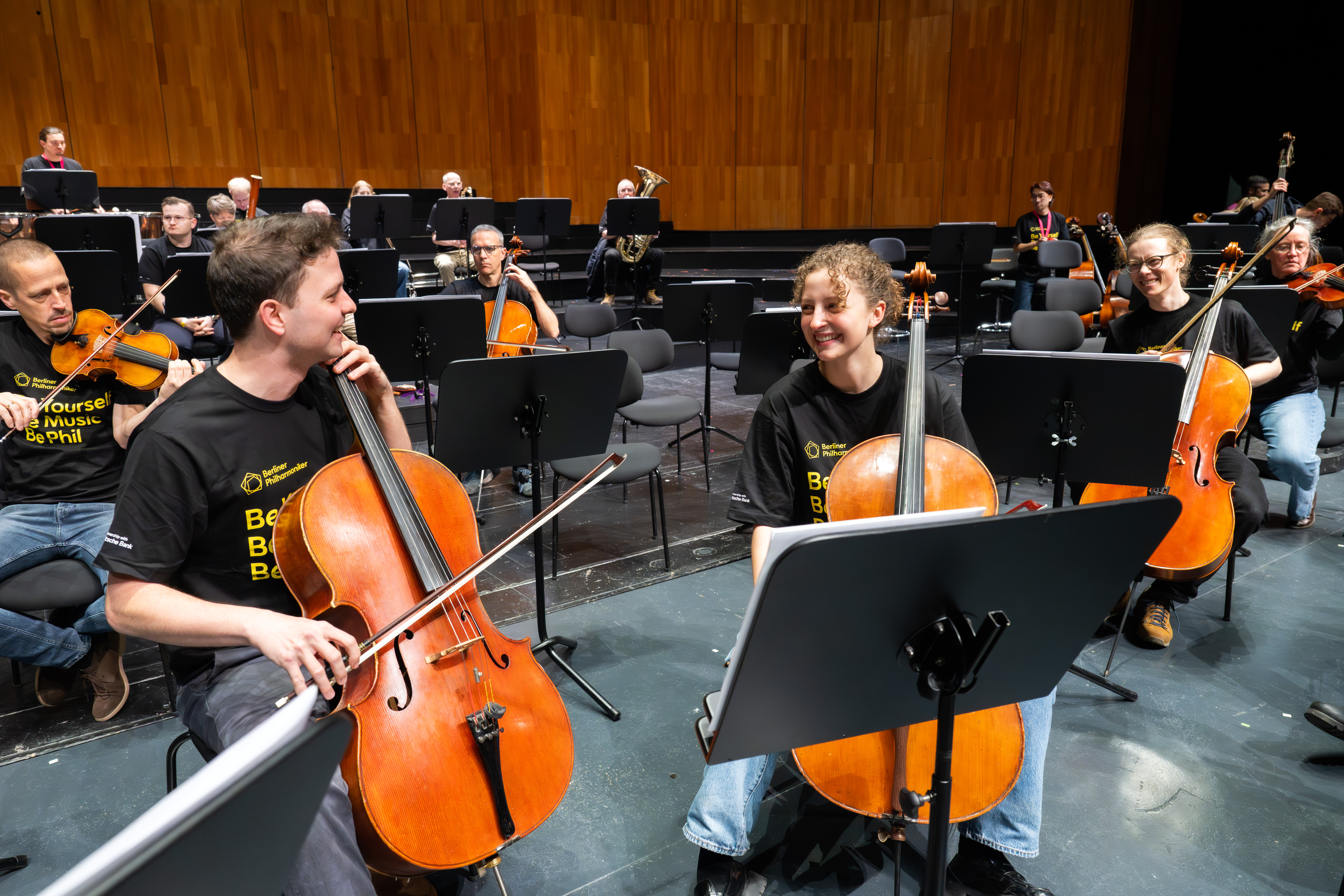 Musiker mit Celli lächeln und unterhalten sich, während sie auf der Bühne sitzen und passende schwarze Hemden tragen. Andere Orchestermitglieder mit Streichinstrumenten sind im Hintergrund an einer Holzwand zu sehen.