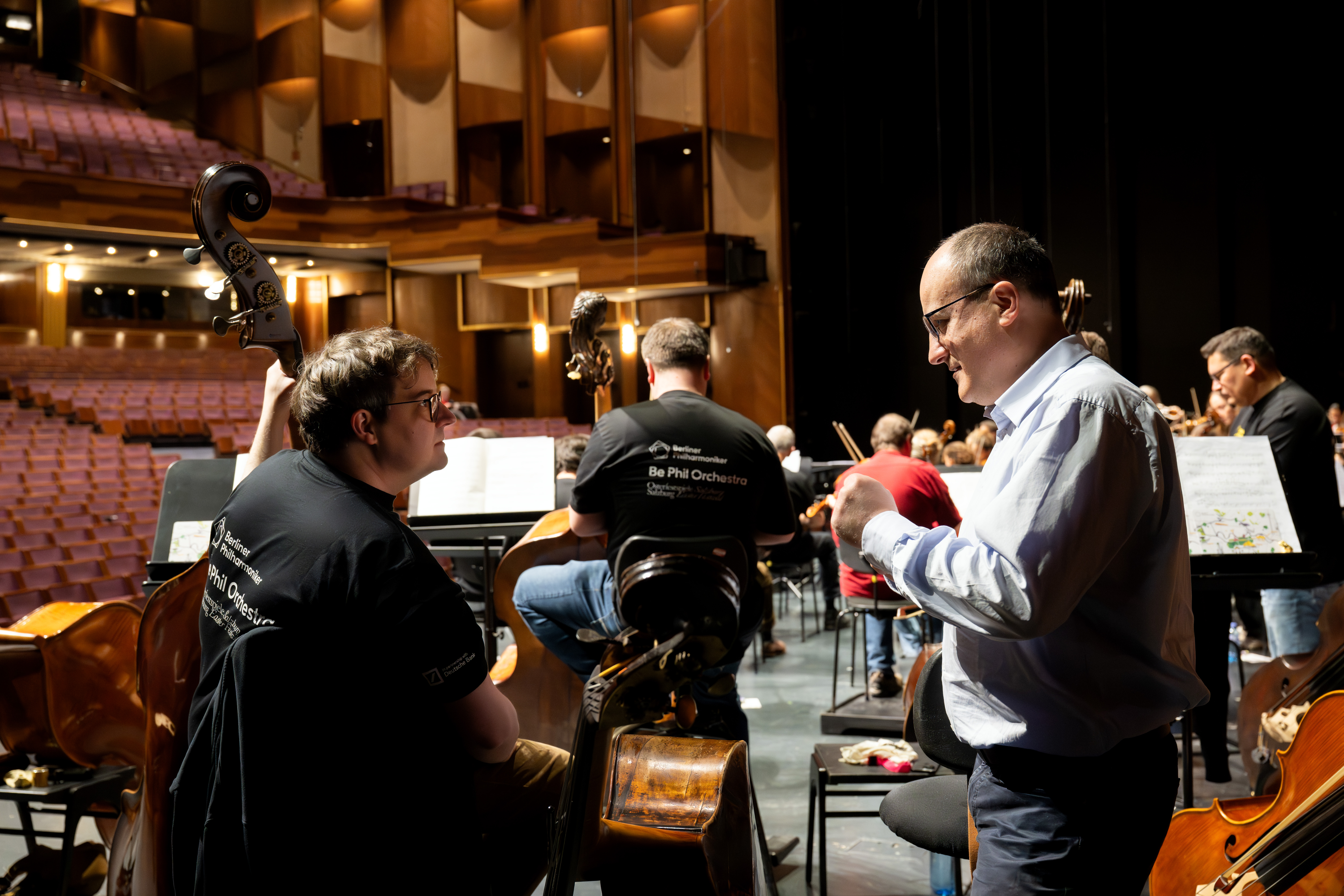 Ein Musiker spricht mit einem Kontrabassisten auf der Bühne während einer Orchesterprobe in einem Konzertsaal, während im Hintergrund andere Musiker und leere Zuschauerplätze zu sehen sind.