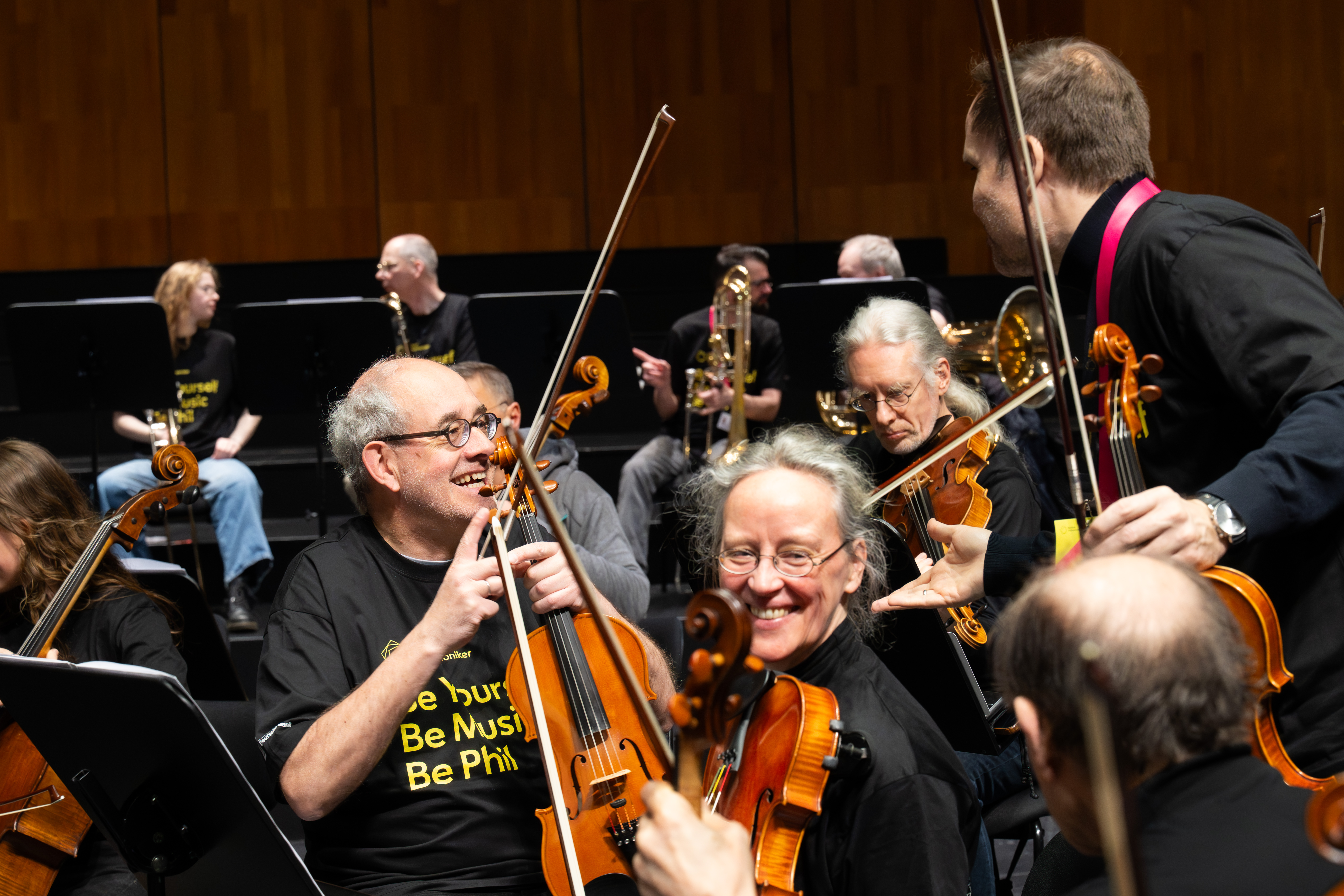Mehrere Orchestermusiker mit Streichinstrumenten lächeln und interagieren spielerisch auf der Bühne, sie tragen passende schwarze Hemden mit gelber Aufschrift; im Hintergrund sind Holzverkleidungen und andere Musiker zu sehen.