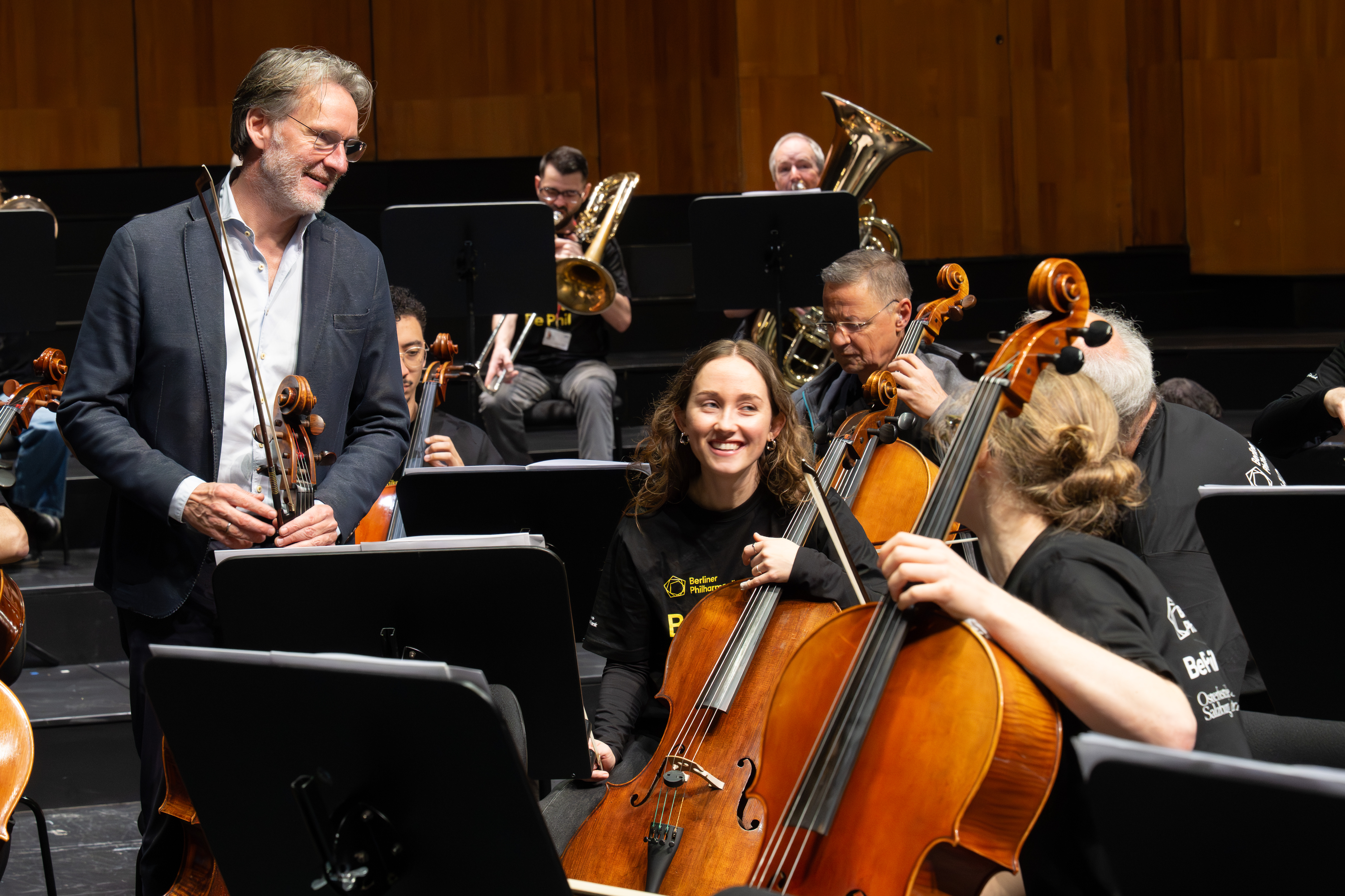 Ein Musiker mit einem Cello lächelt den Cellisten während einer Orchesterprobe zu. Musiker spielen verschiedene Instrumente in einem Konzertsaal mit Holzvertäfelung im Hintergrund.