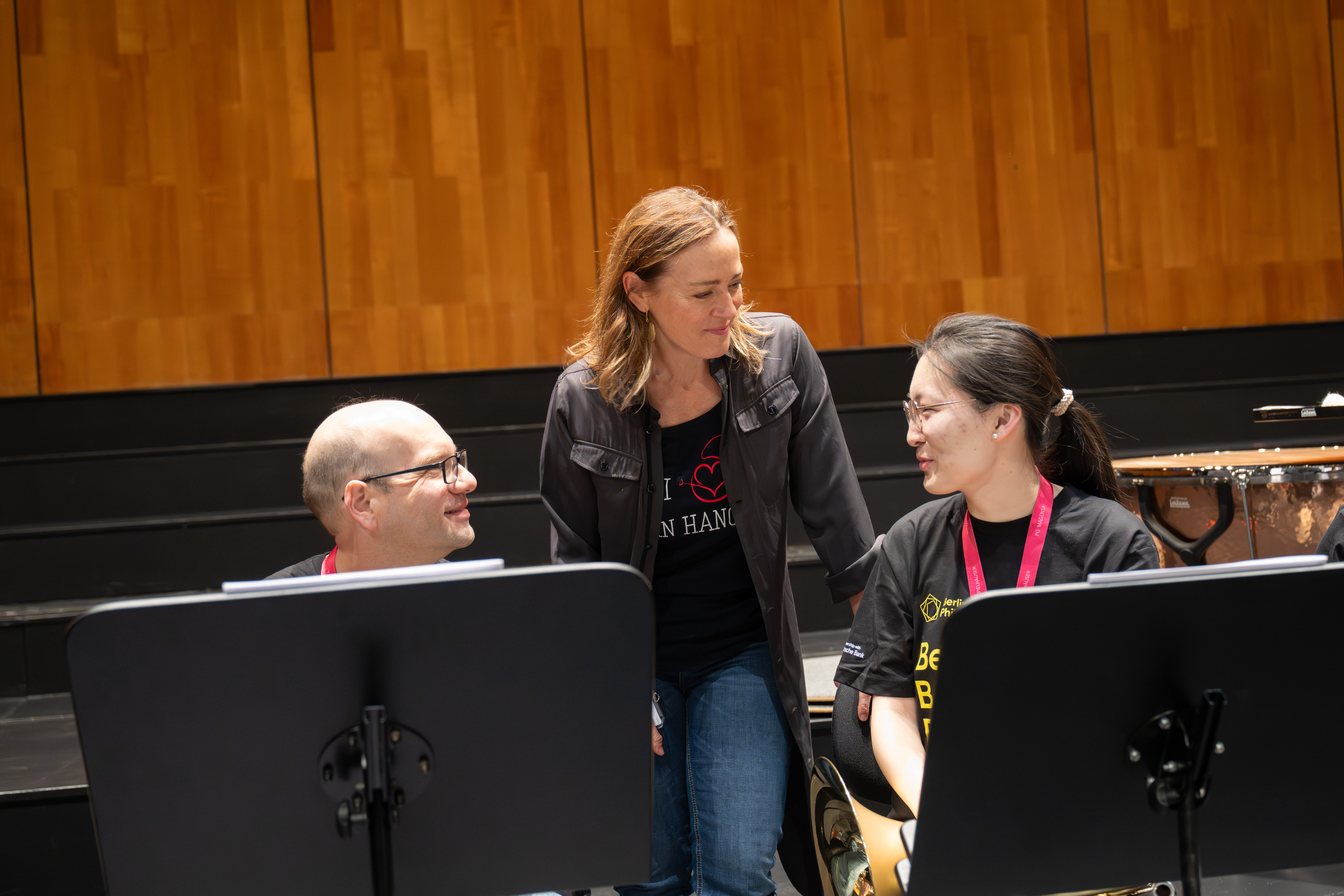 Drei Musiker auf einer Bühne mit Notenständern; zwei sitzen und halten Instrumente, während einer zwischen ihnen steht, lächelt und spricht. Im Hintergrund sind eine Holzwand und Schlaginstrumente zu sehen.