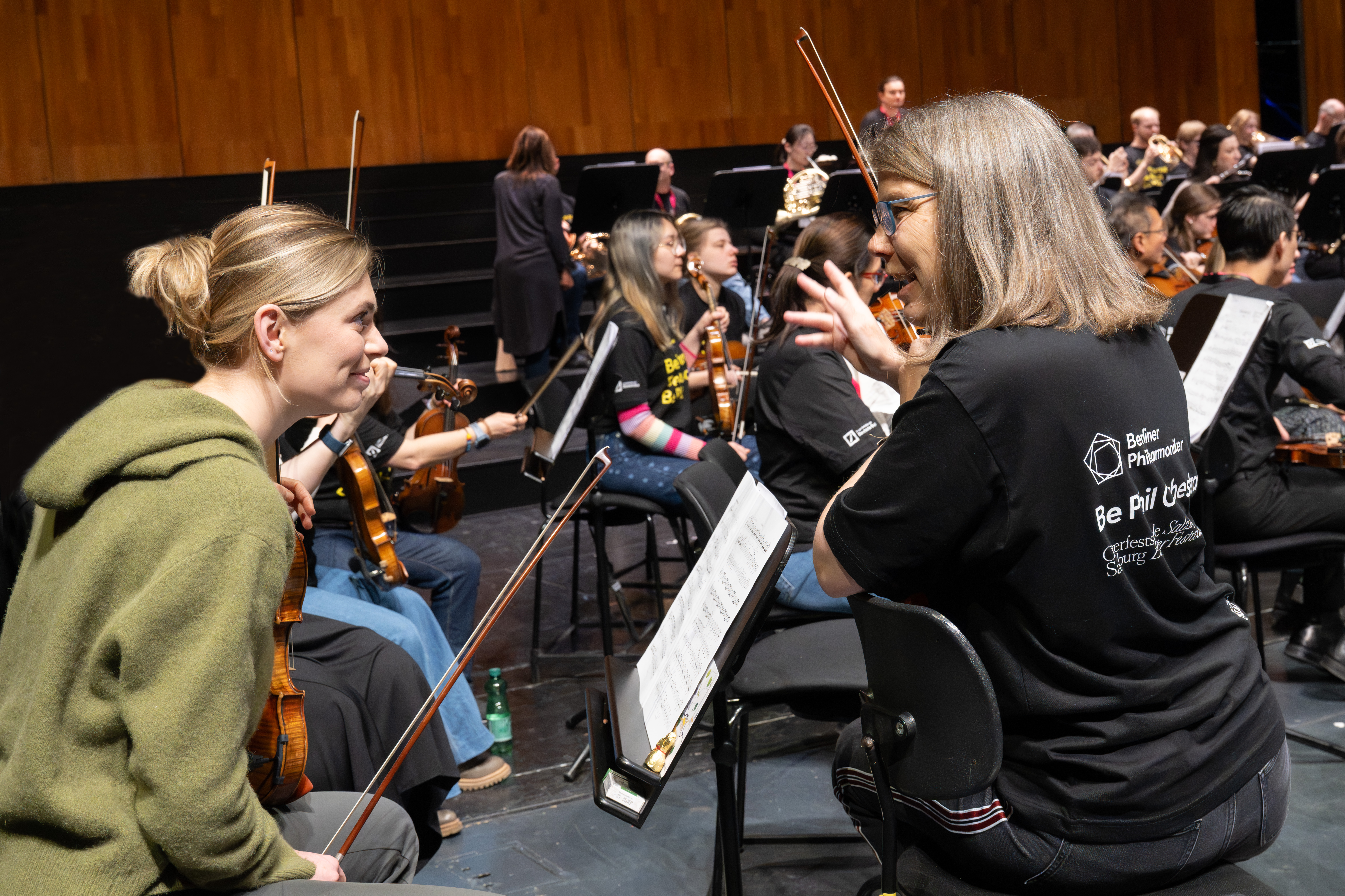 Zwei Frauen mit Geigen sitzen auf der Bühne und unterhalten sich während einer Orchesterprobe. Im Hintergrund sind andere Musiker und Notenständer zu sehen. Eine Frau gestikuliert mit ihrer Hand, während sie sich unterhalten.