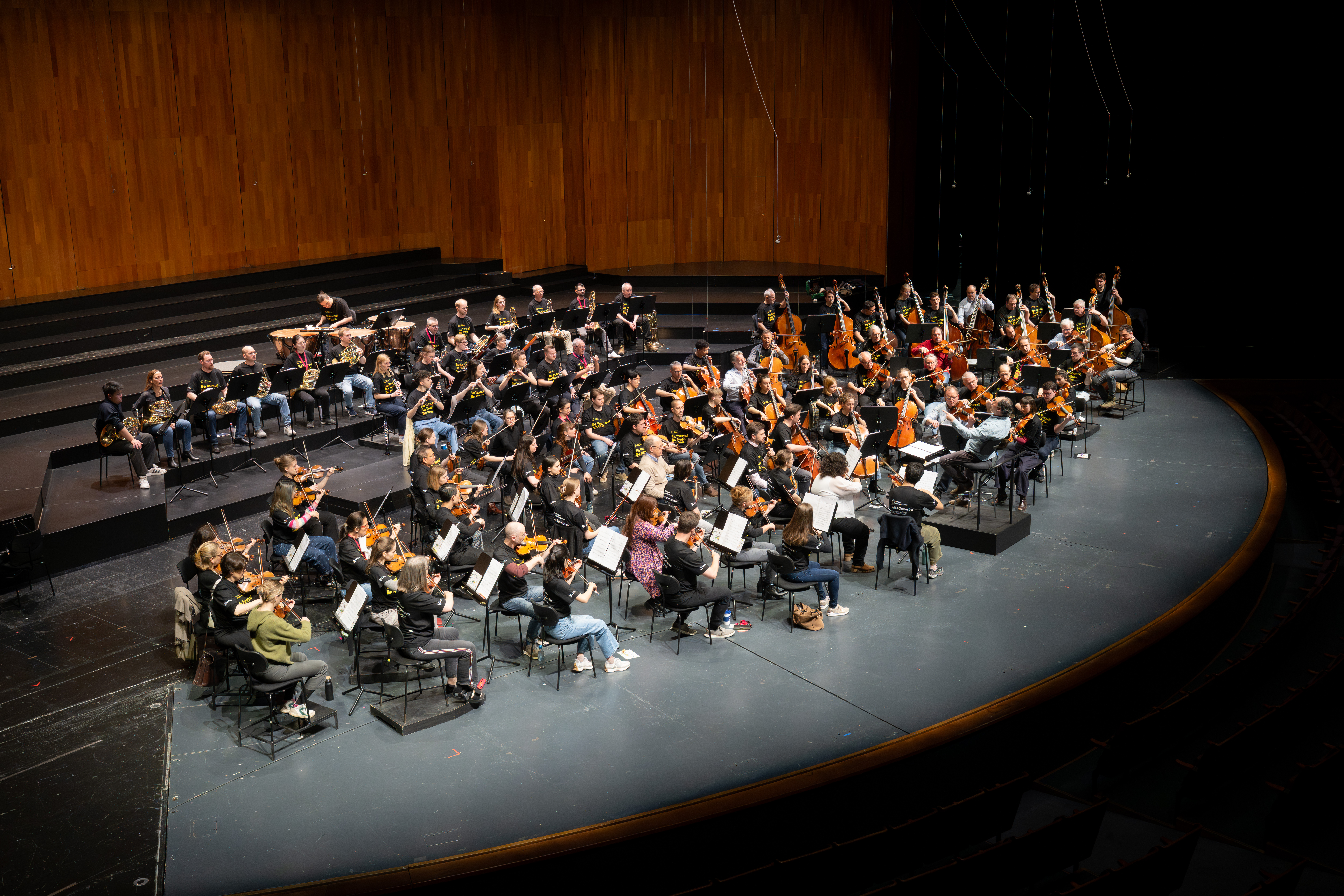 Ein großes Orchester probt auf der Bühne, wobei die Musiker verschiedene Instrumente spielen, die in Abschnitten angeordnet sind. Im Hintergrund sind Holzverkleidungen und leere Sitzreihen hinter den Musikern zu sehen.