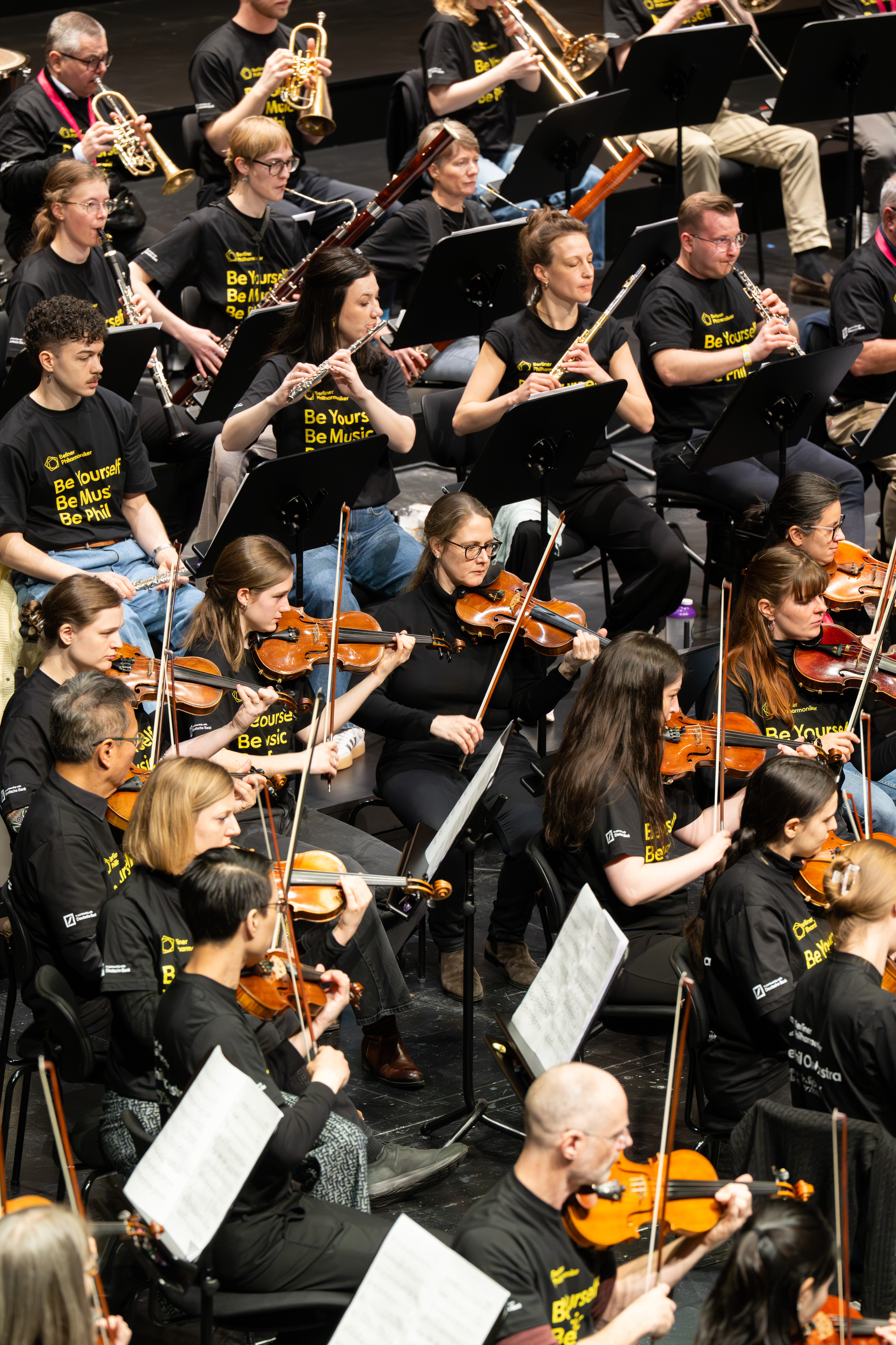 Ein großes Orchester von Musikern in passenden schwarzen Hemden spielt auf der Bühne Streich- und Blasinstrumente. Notenständer stehen vor ihnen, und auf den Hemden steht »Be Yourself. Be Music. Be Phil.«.
