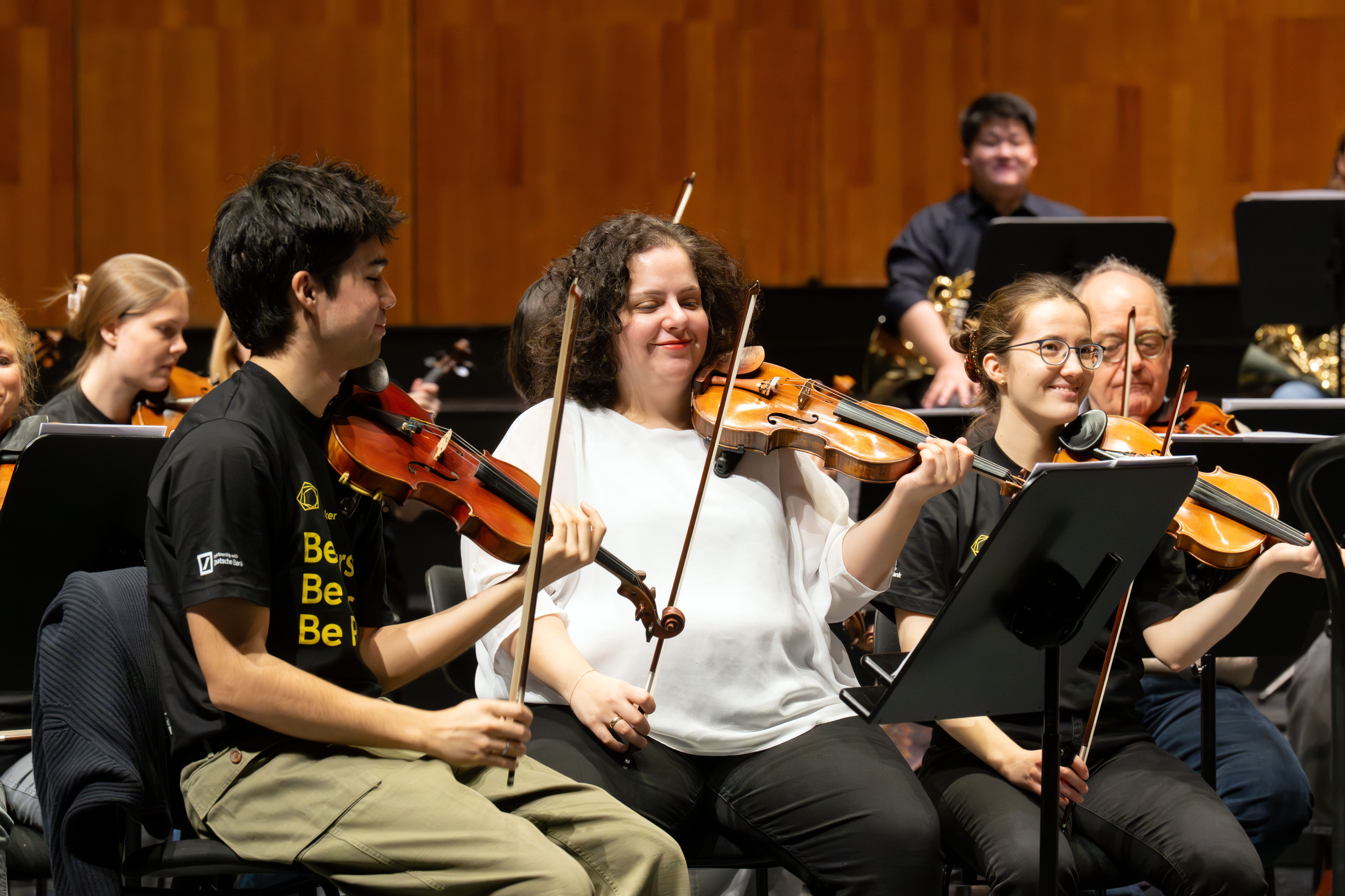 Eine Gruppe von Musikern eines Orchesters spielt Geigen und lächelt während einer Probe; sie sitzen mit Notenständern, tragen legere Kleidung und haben eine Holzwand im Hintergrund.