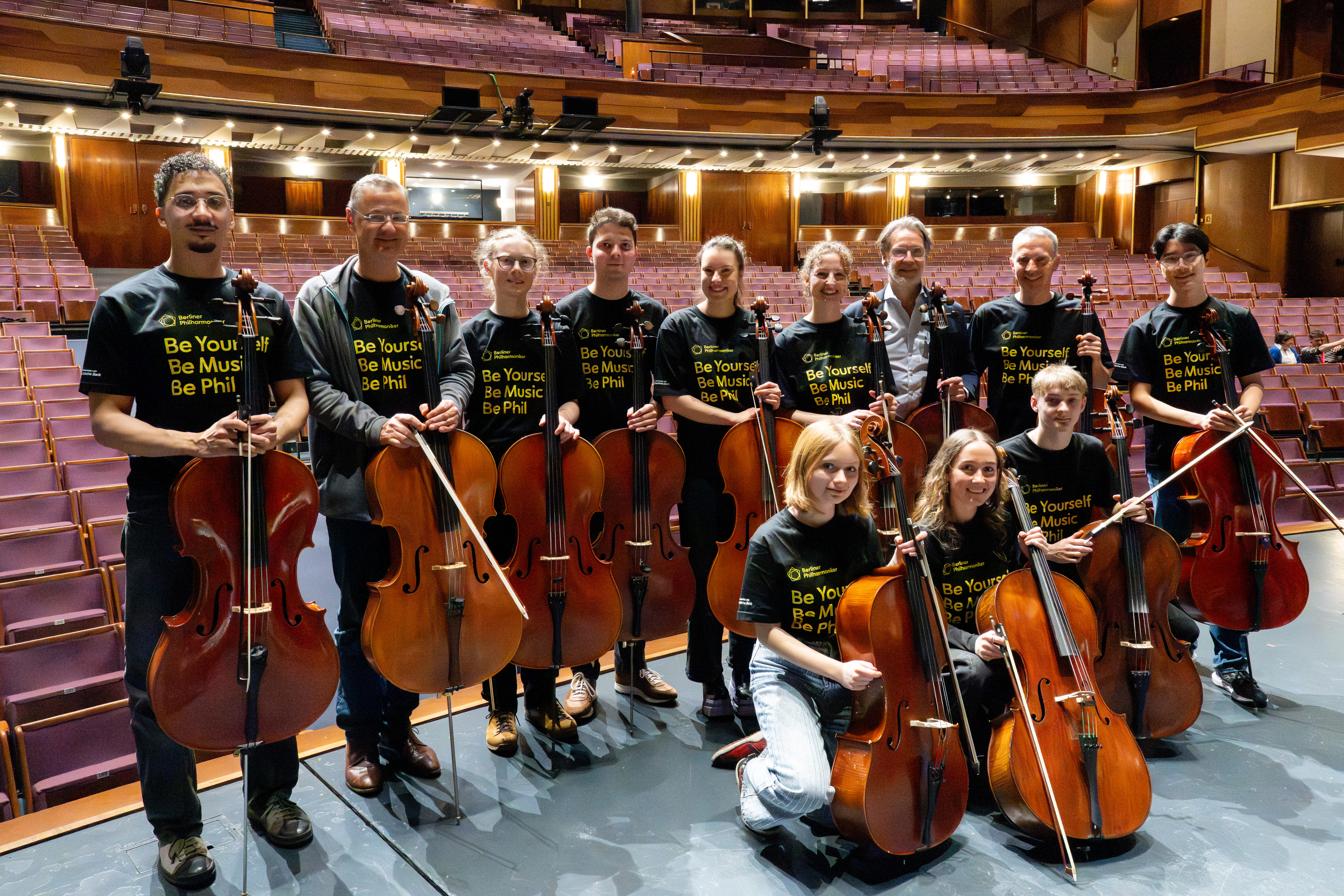 Elf Personen mit Cellos stehen auf einer Theaterbühne und tragen passende »Be Yourself, Be Music, Be Phil«-Shirts, während im Hintergrund leere Sitzreihen und der Balkon zu sehen sind.