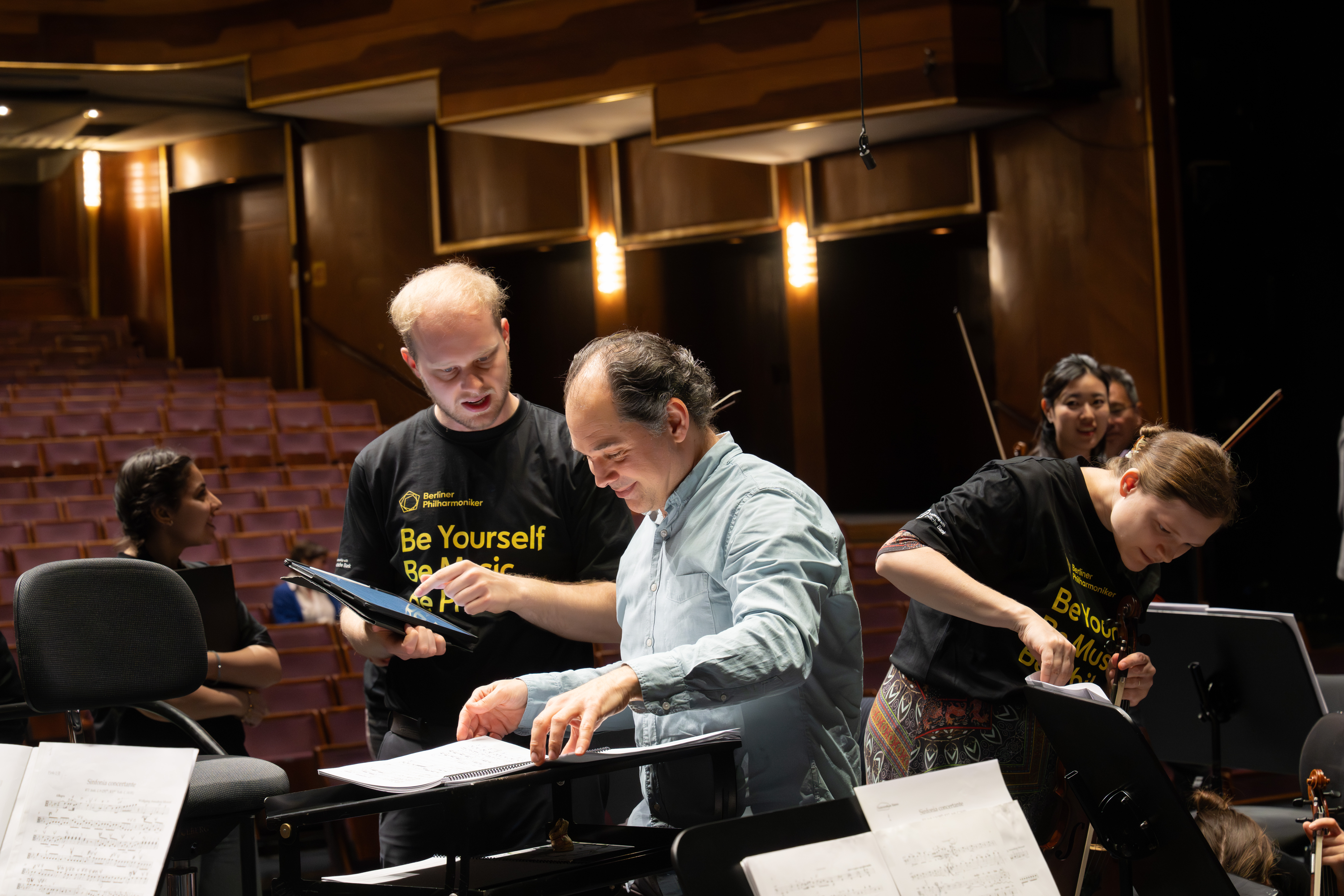 Ein Dirigent bespricht mit einem jungen Musiker auf der Bühne die Noten, während andere in passenden »Be Yourself, Be Music«-Shirts ihre Instrumente in einem Konzertsaal mit leeren Sitzen vorbereiten.