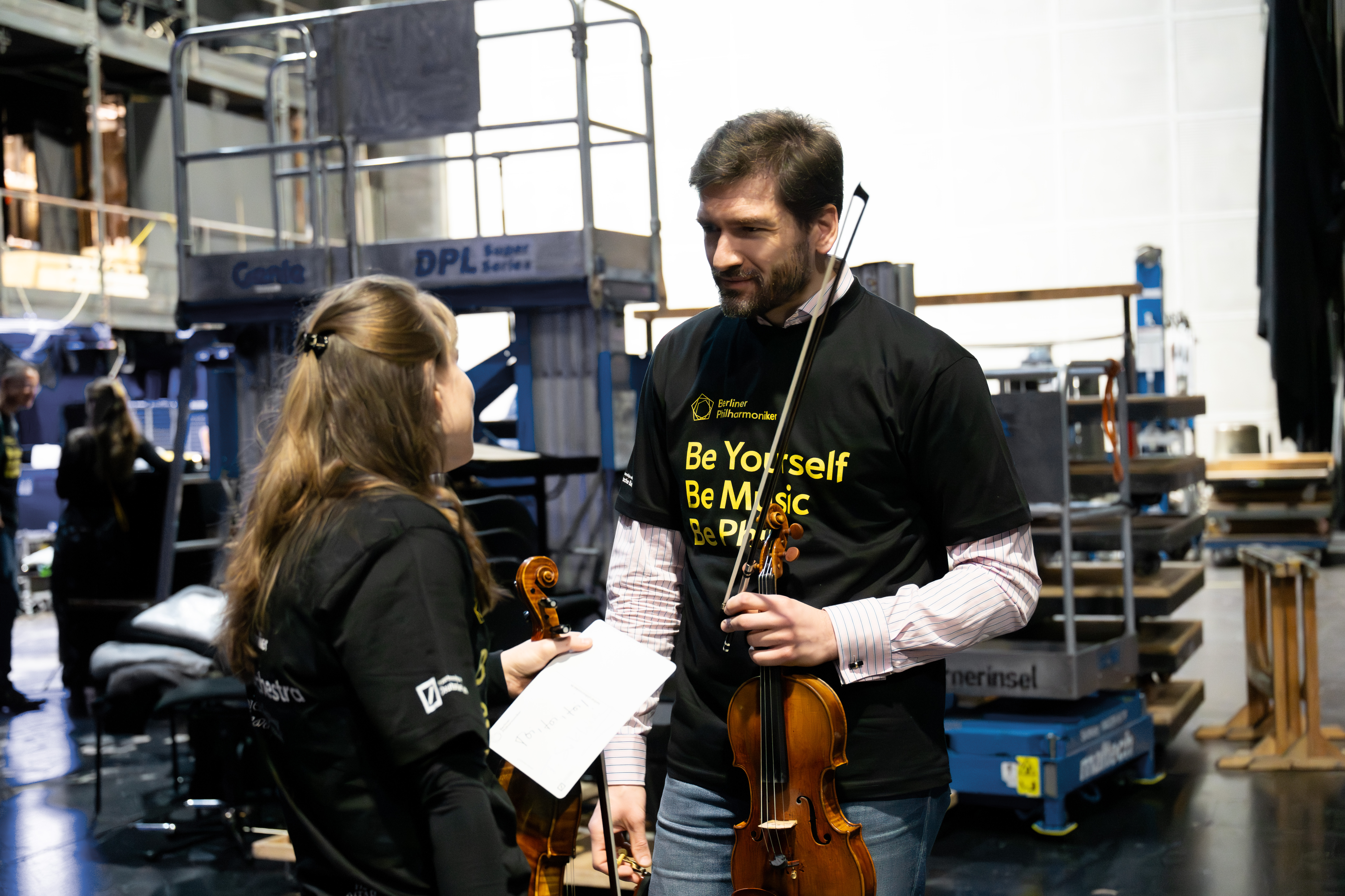 Zwei Musiker mit »Be Yourself, Be Music, Be Phil«-Shirts, die Geigen halten und sich miteinander unterhalten; im Hintergrund sind Bühnentechnik und Regale zu sehen.