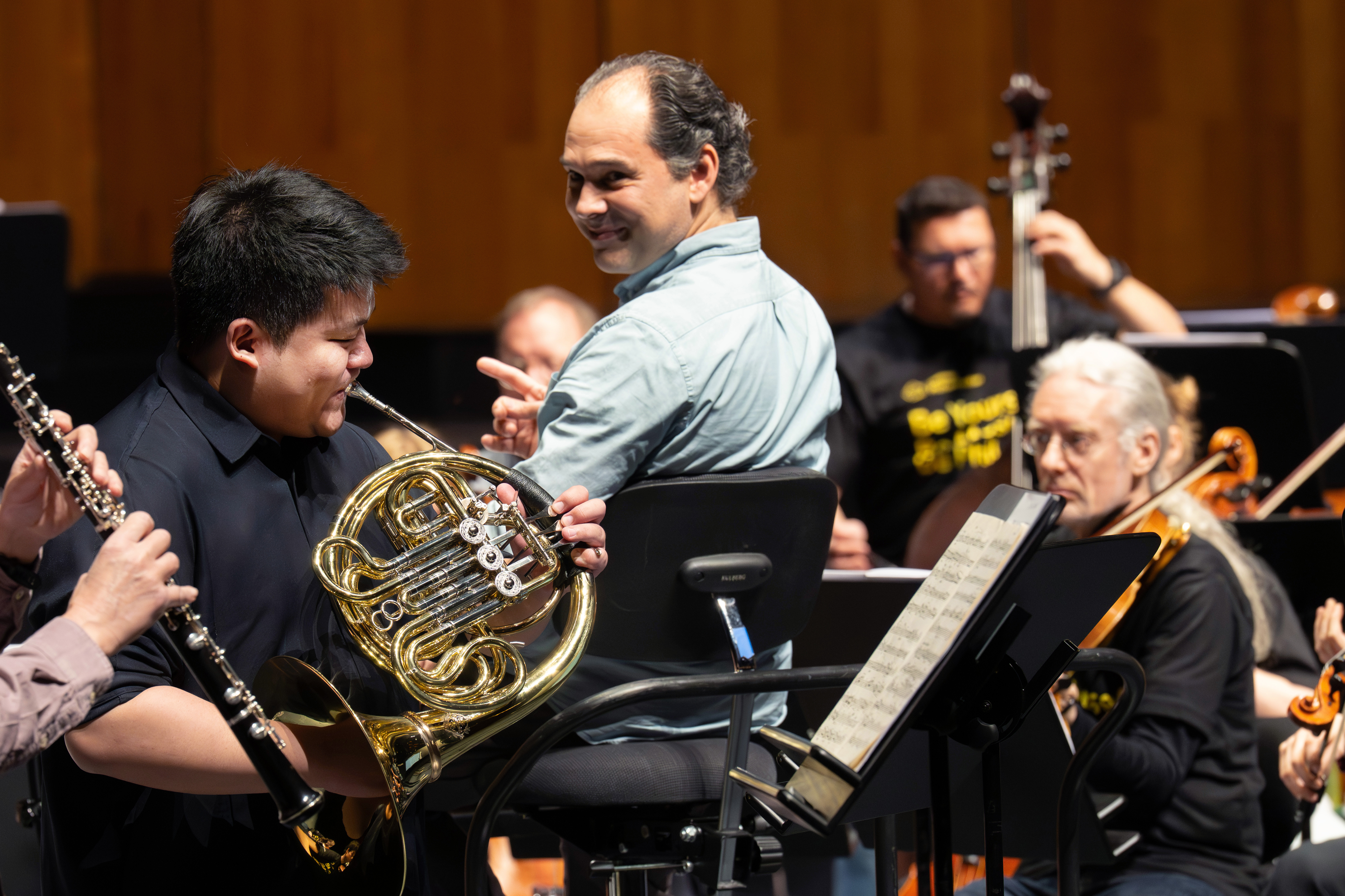 Ein Dirigent lächelt und gestikuliert in Richtung eines Waldhornspielers während einer Orchesterprobe. Musiker mit verschiedenen Instrumenten erscheinen konzentriert, im Vordergrund sind Notenblätter zu sehen.