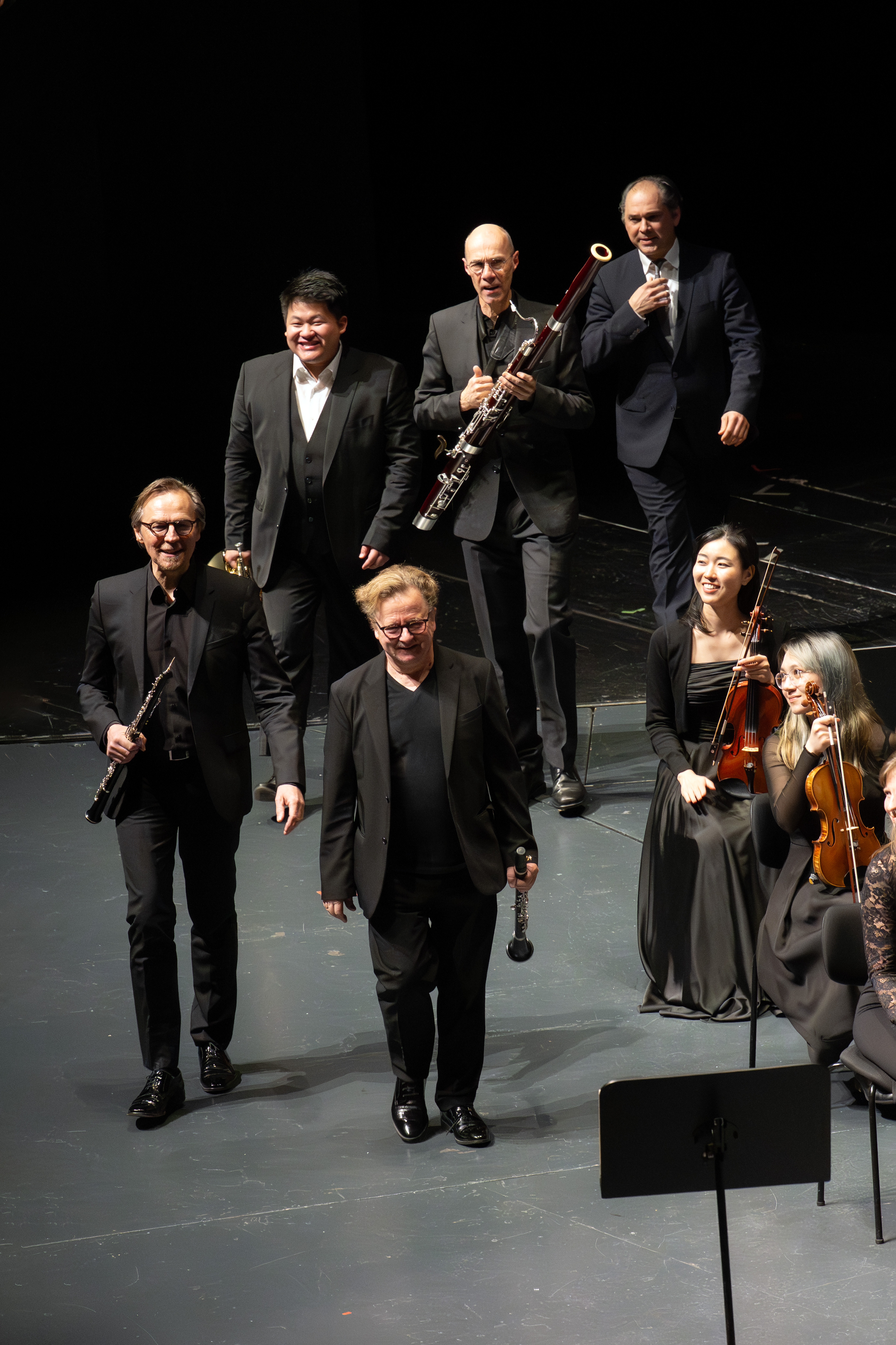 Sechs Musiker, fünf im Stehen und zwei im Sitzen mit Streichinstrumenten, betreten lächelnd und in formeller schwarzer Kleidung die Bühne und scheinen den Applaus nach einem Auftritt zu quittieren.
