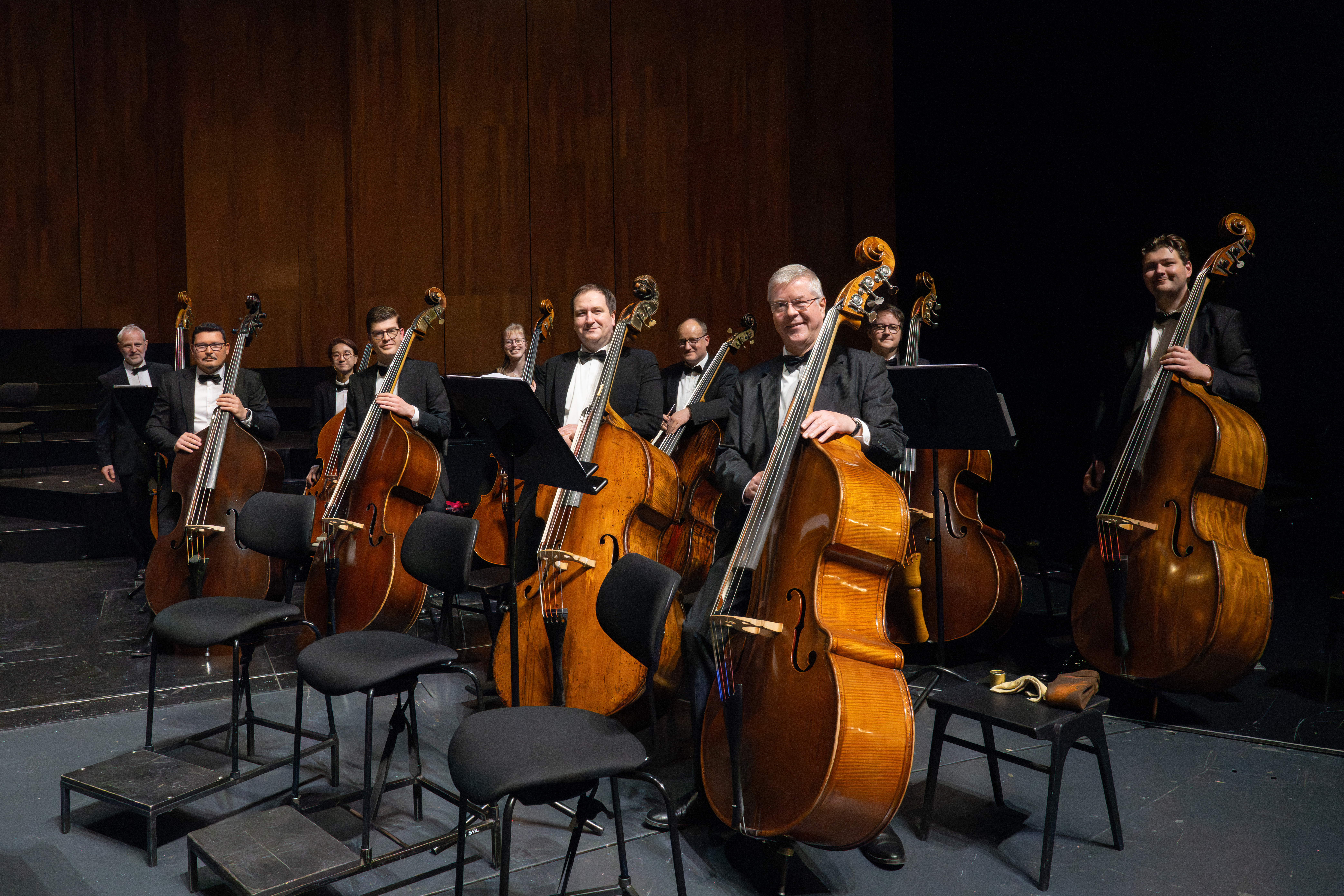 Neun Musiker in formeller Kleidung stehen mit Kontrabässen auf der Bühne und lächeln in die Kamera. Vor ihnen sind Notenständer und leere Stühle aufgestellt, dahinter ein dunkler Hintergrund.