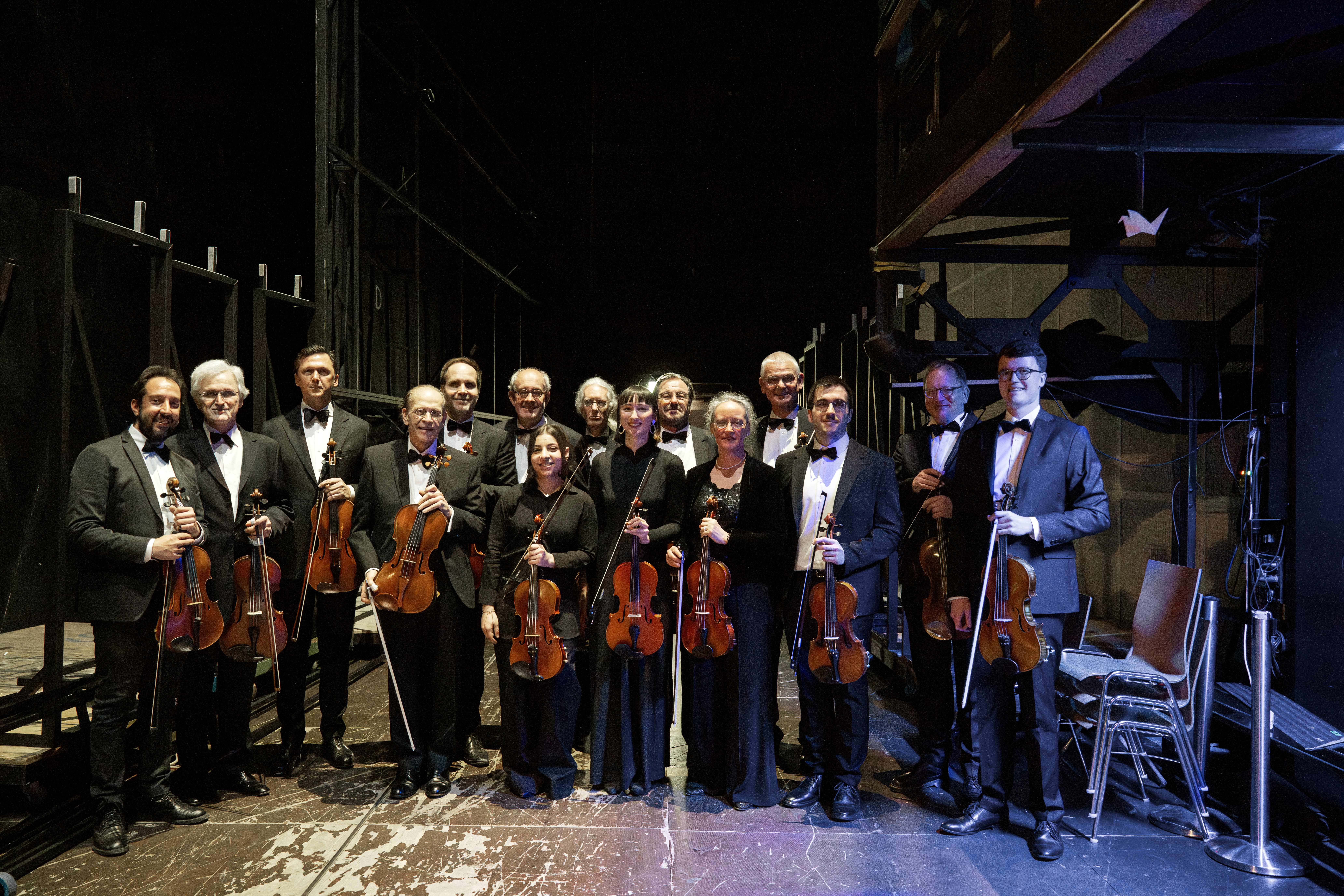 Eine Gruppe von dreizehn förmlich gekleideten Musikern mit Bratschen steht hinter der Bühne auf einer schwach beleuchteten Bühne zusammen und lächelt in die Kamera.