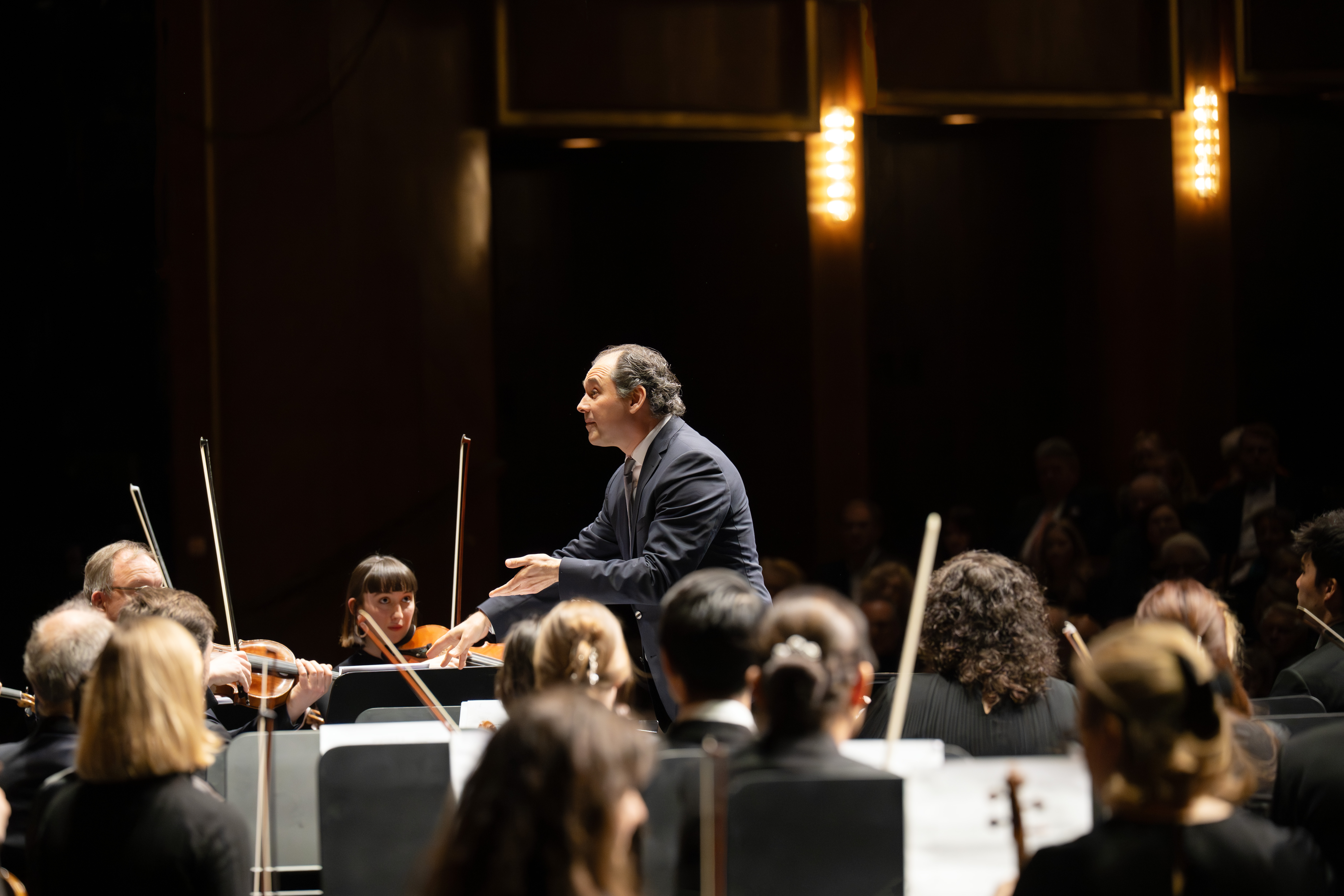 Ein Dirigent in einem dunklen Anzug leitet ein Orchester während einer Live-Aufführung, während die Musiker Streichinstrumente spielen und das Publikum im schwach beleuchteten Hintergrund zu sehen ist.