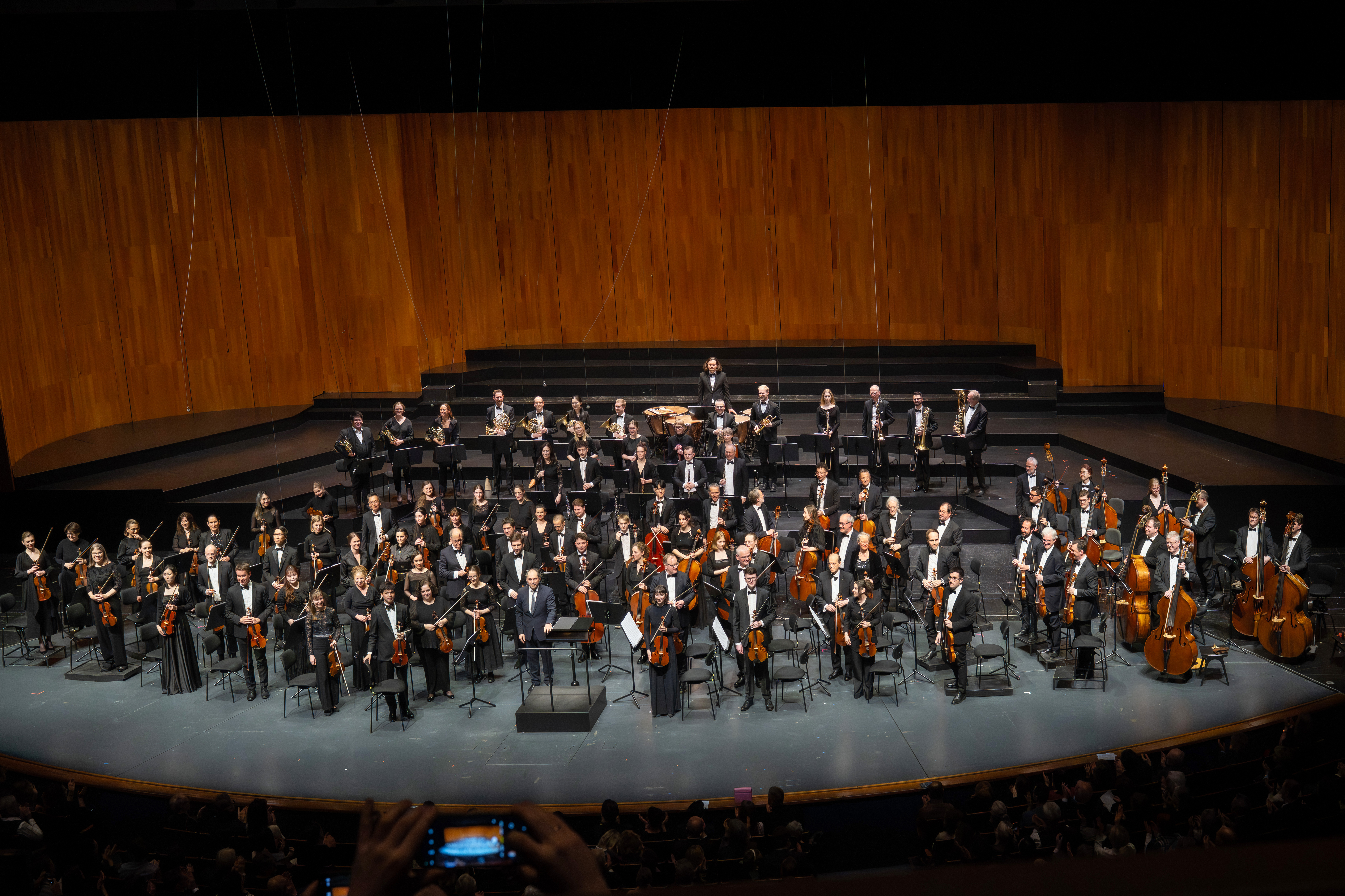 Ein Orchester steht mit seinen Instrumenten auf der Bühne und blickt auf das Publikum in einem modernen Konzertsaal mit einem Holzhintergrund. Die Musiker tragen formelle schwarze Kleidung und im Publikum sitzen Menschen.