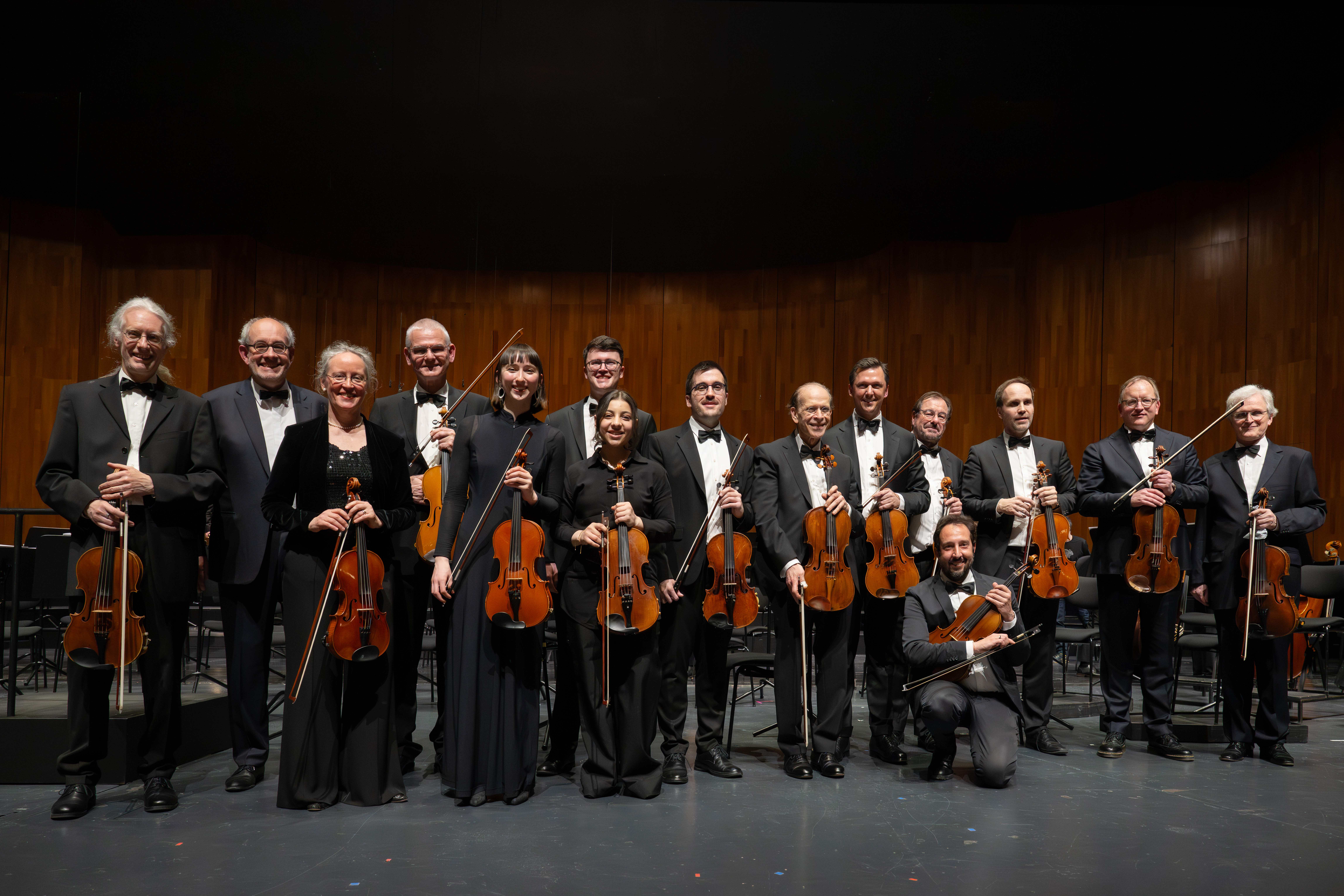 Eine Gruppe von dreizehn Musikern in formeller schwarzer Kleidung posiert auf der Bühne mit Bratschen vor einem hölzernen Hintergrund.