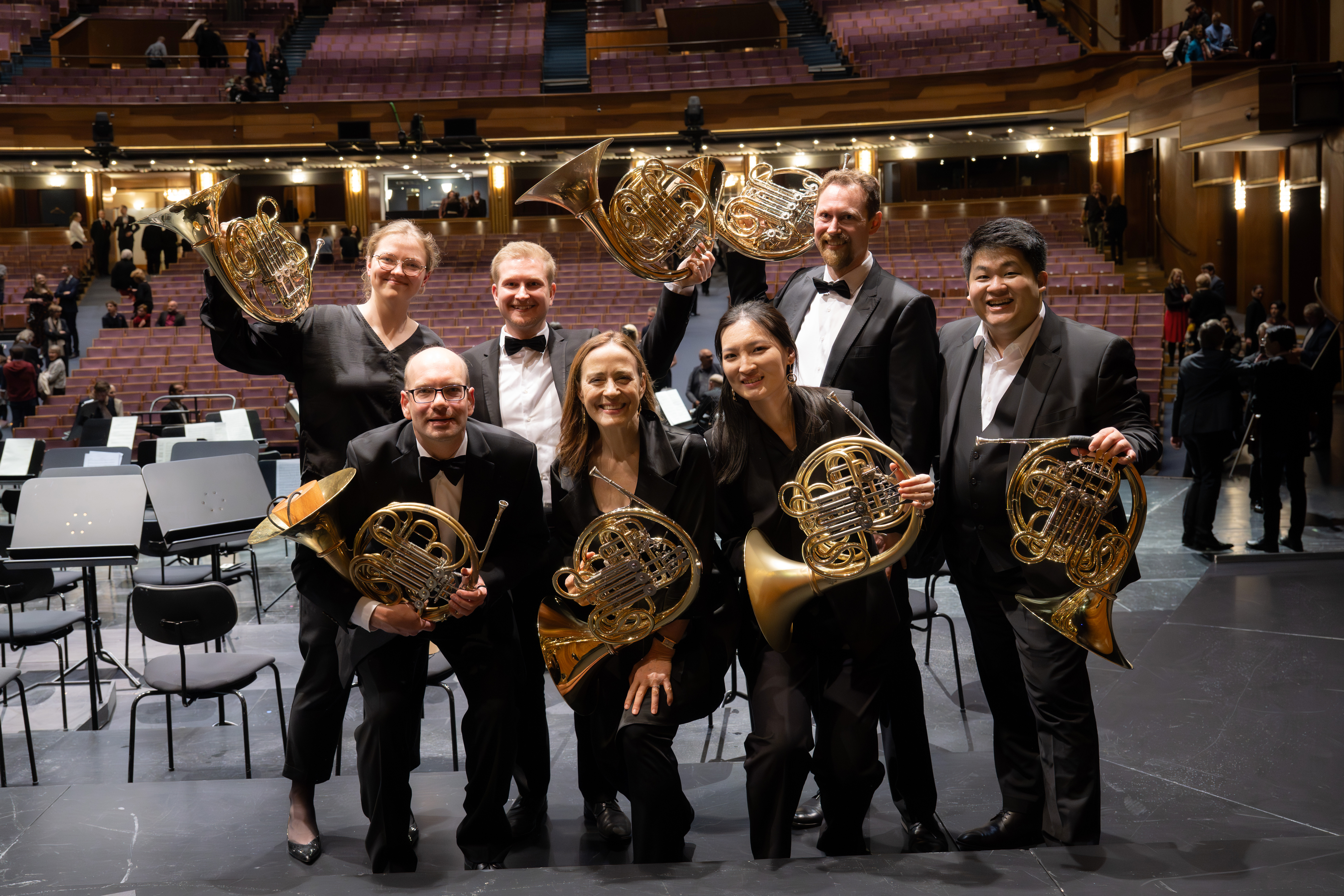 Sieben Musiker in formeller schwarzer Kleidung posieren auf der Bühne eines Konzertsaals, lächeln und halten Waldhörner. Im Hintergrund sind Reihen von leeren Sitzen und andere Musiker zu sehen.