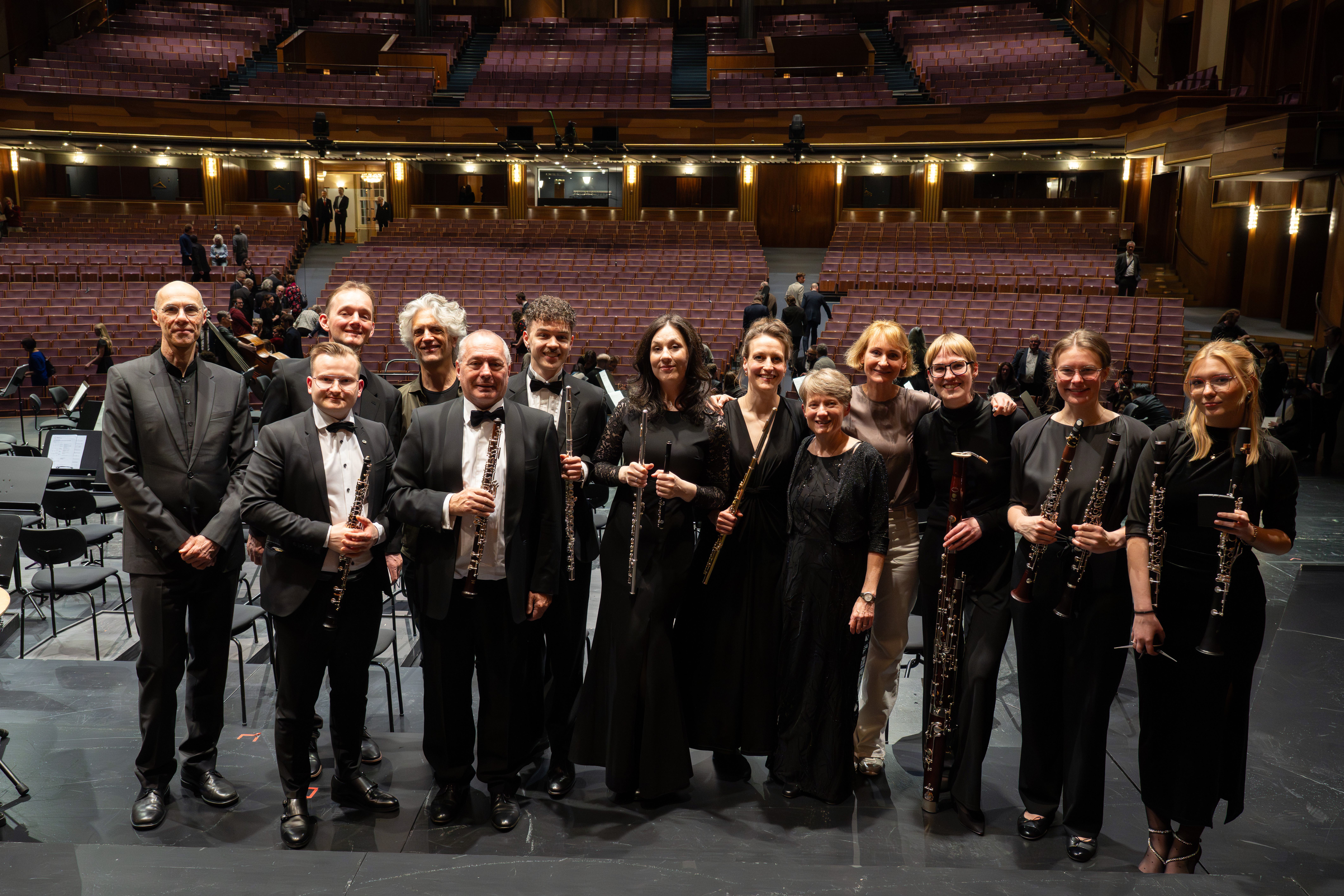 Eine Gruppe von formell gekleideten Musikern mit Holzblasinstrumenten steht lächelnd auf der Bühne eines Konzertsaals, mit leeren Sitzen und ein paar Leuten im Hintergrund.