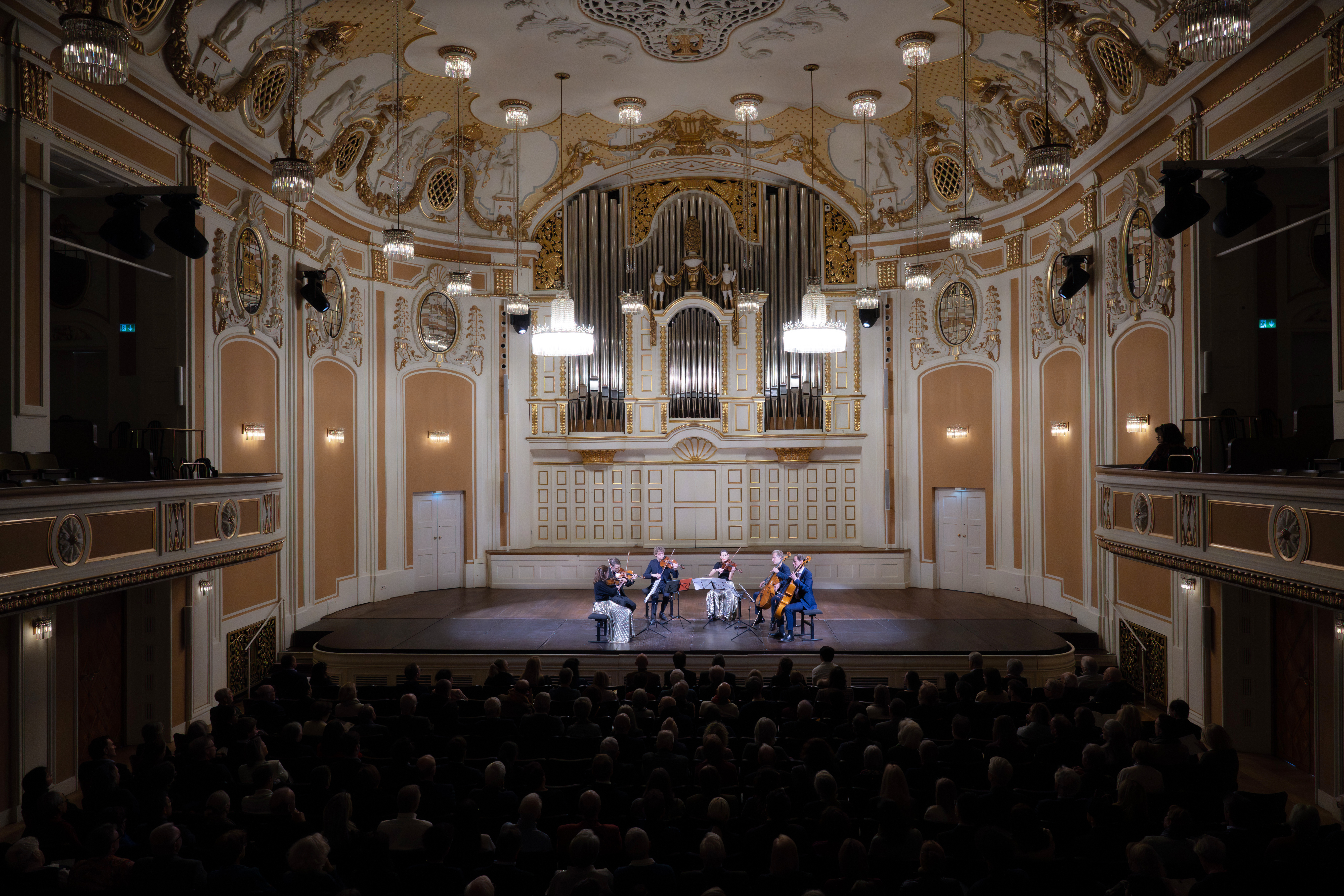 Ein Streichquartett spielt auf der Bühne eines mit Gold verzierten Konzertsaals mit einer großen Pfeifenorgel und einem davor sitzenden Publikum.