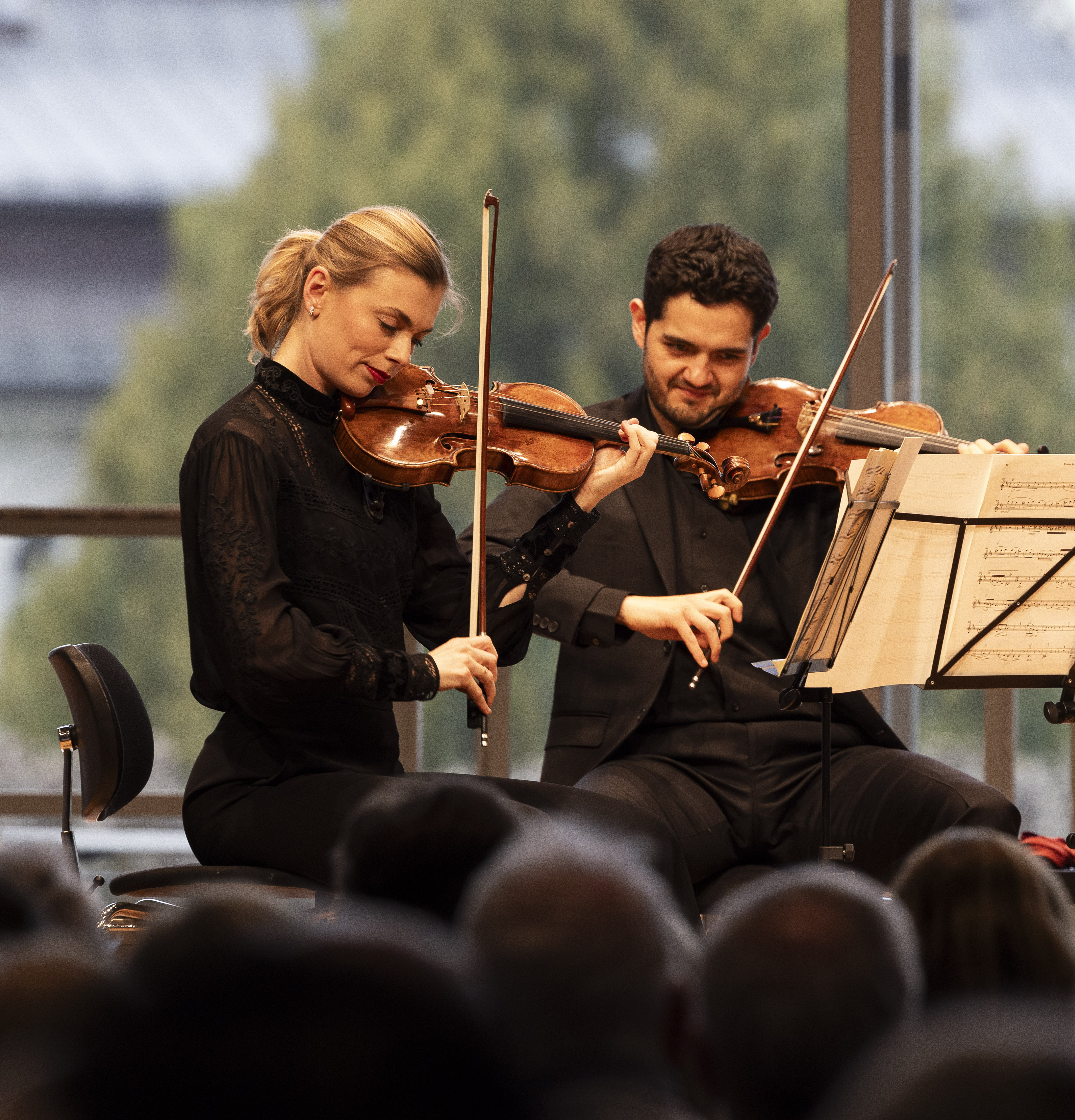 Zwei Geiger, eine Frau und ein Mann, spielen gemeinsam auf der Bühne und lesen Noten. Sie tragen formelle schwarze Kleidung, ein Publikum schaut zu und im Hintergrund sind große Fenster mit Bäumen zu sehen.