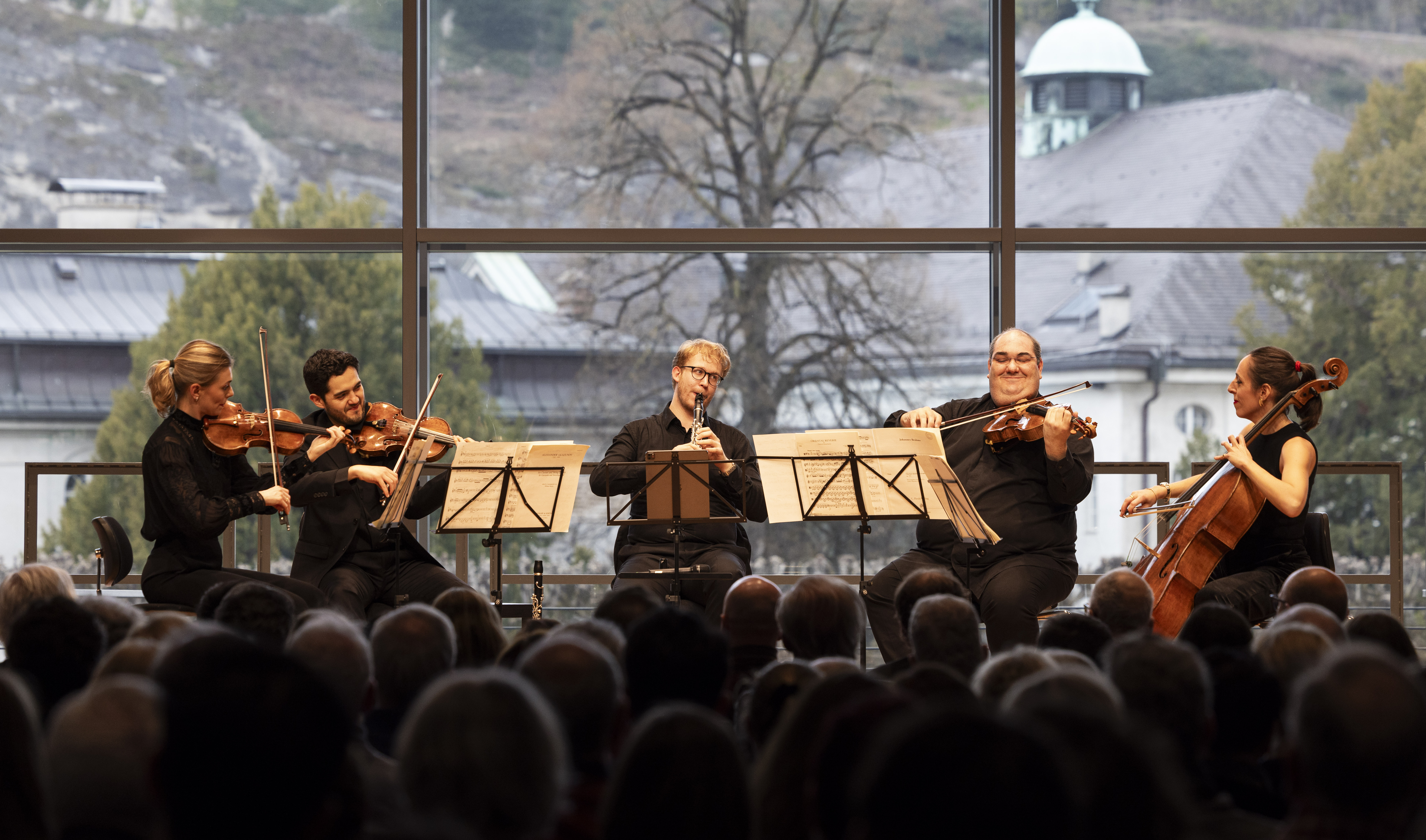 Fünf Musiker spielen auf der Bühne ein Kammermusikkonzert mit Streichern und Holzblasinstrumenten, während das Publikum zuschaut und große Fenster einen malerischen, baumbestandenen Hintergrund zeigen.