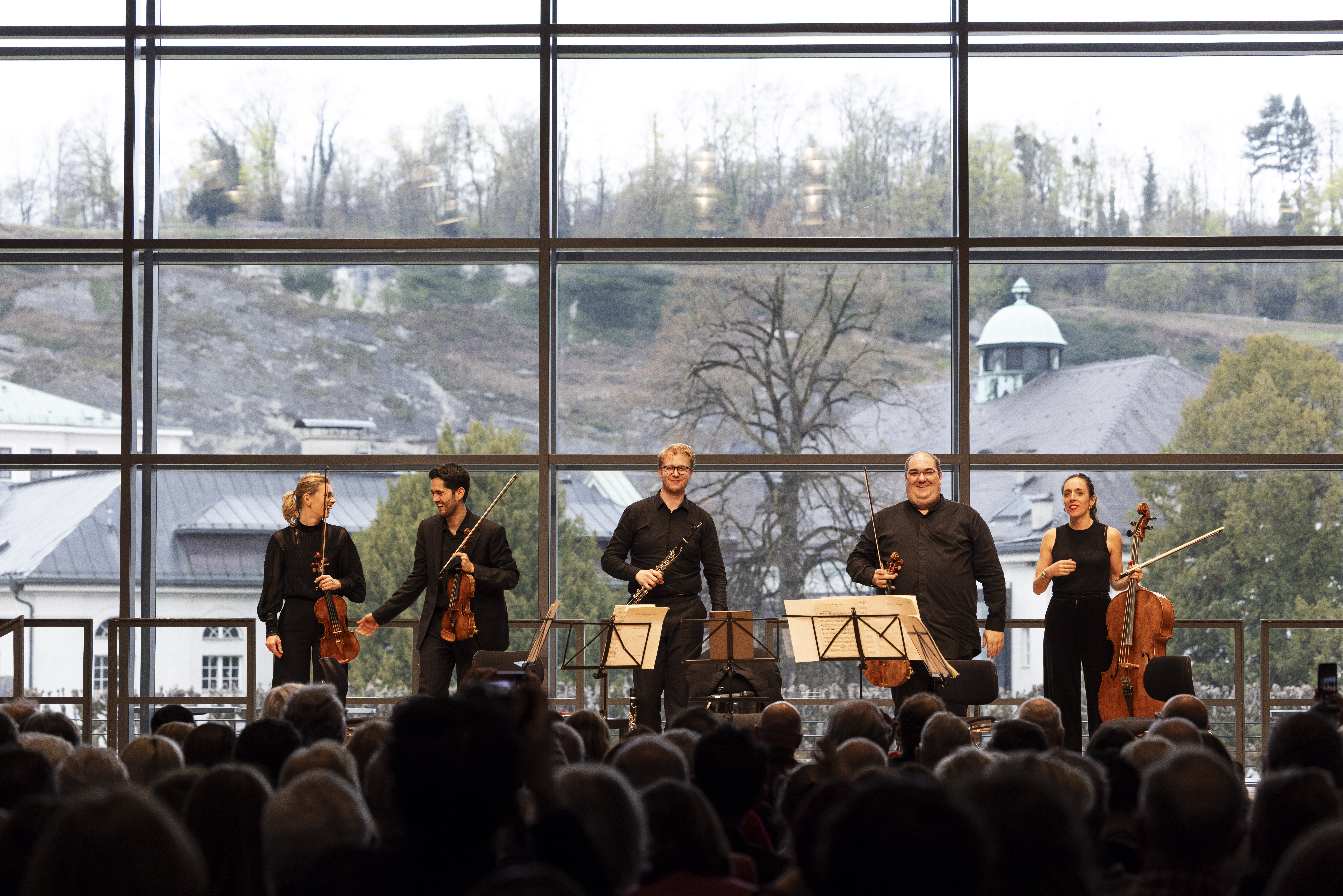 Fünf Musiker mit Streichinstrumenten stehen auf der Bühne, lächeln und verbeugen sich vor einem Publikum vor einem großen Fenster mit Blick auf Bäume und Gebäude. Auf der Bühne sind Notenständer und Stühle zu sehen.