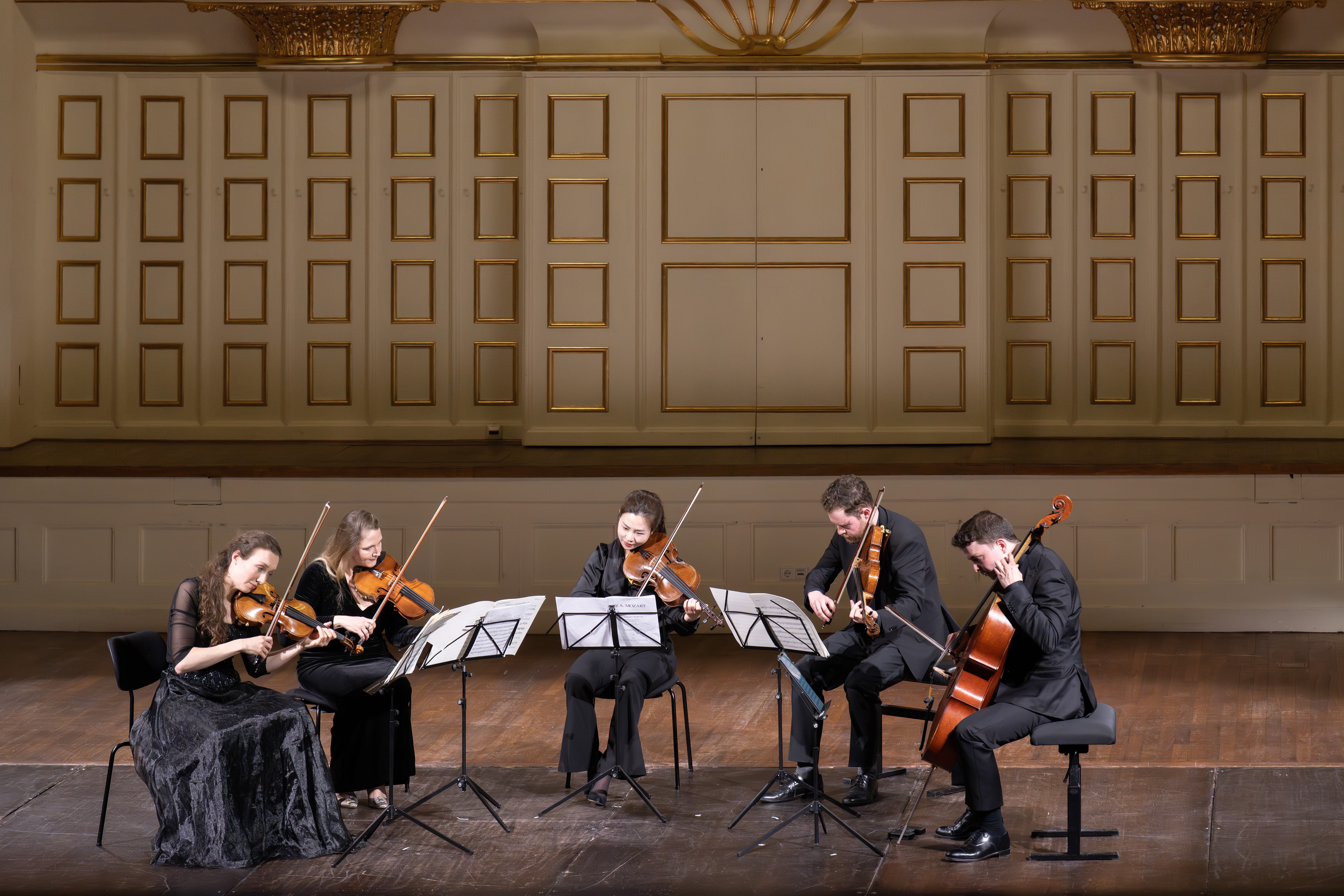 Fünf Musiker in formeller schwarzer Kleidung spielen auf einer hölzernen Bühne auf Streichinstrumenten mit Notenständern vor einer dekorativen Wand mit Goldverzierungen und getäfelten Türen.