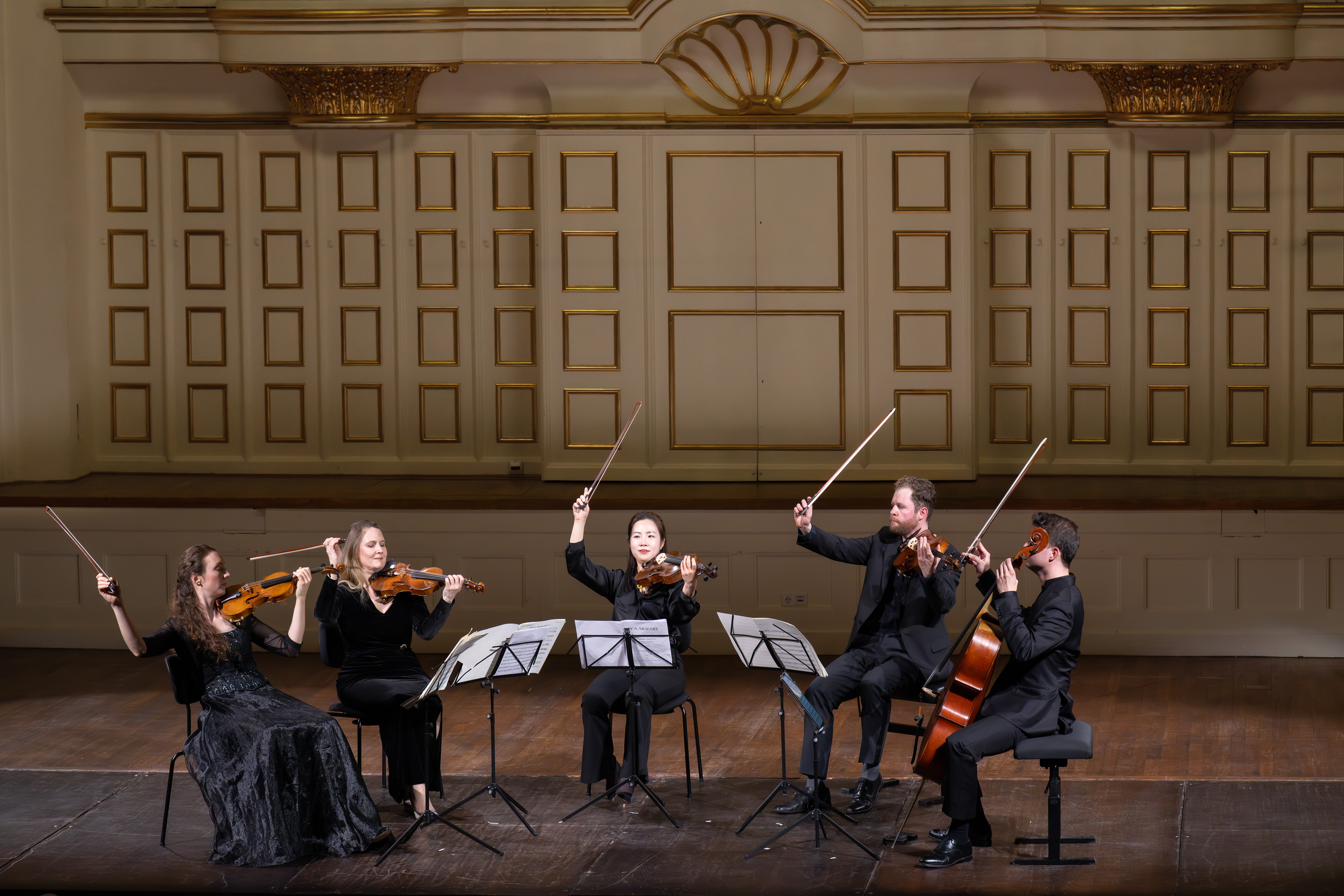 Fünf Musiker in formeller schwarzer Kleidung treten auf der Bühne als Streichquintett auf, jeder spielt ein Streichinstrument mit erhobenen Bögen, sitzend in einem großen Konzertsaal mit verschnörkeltem Gold- und Weißdekor.