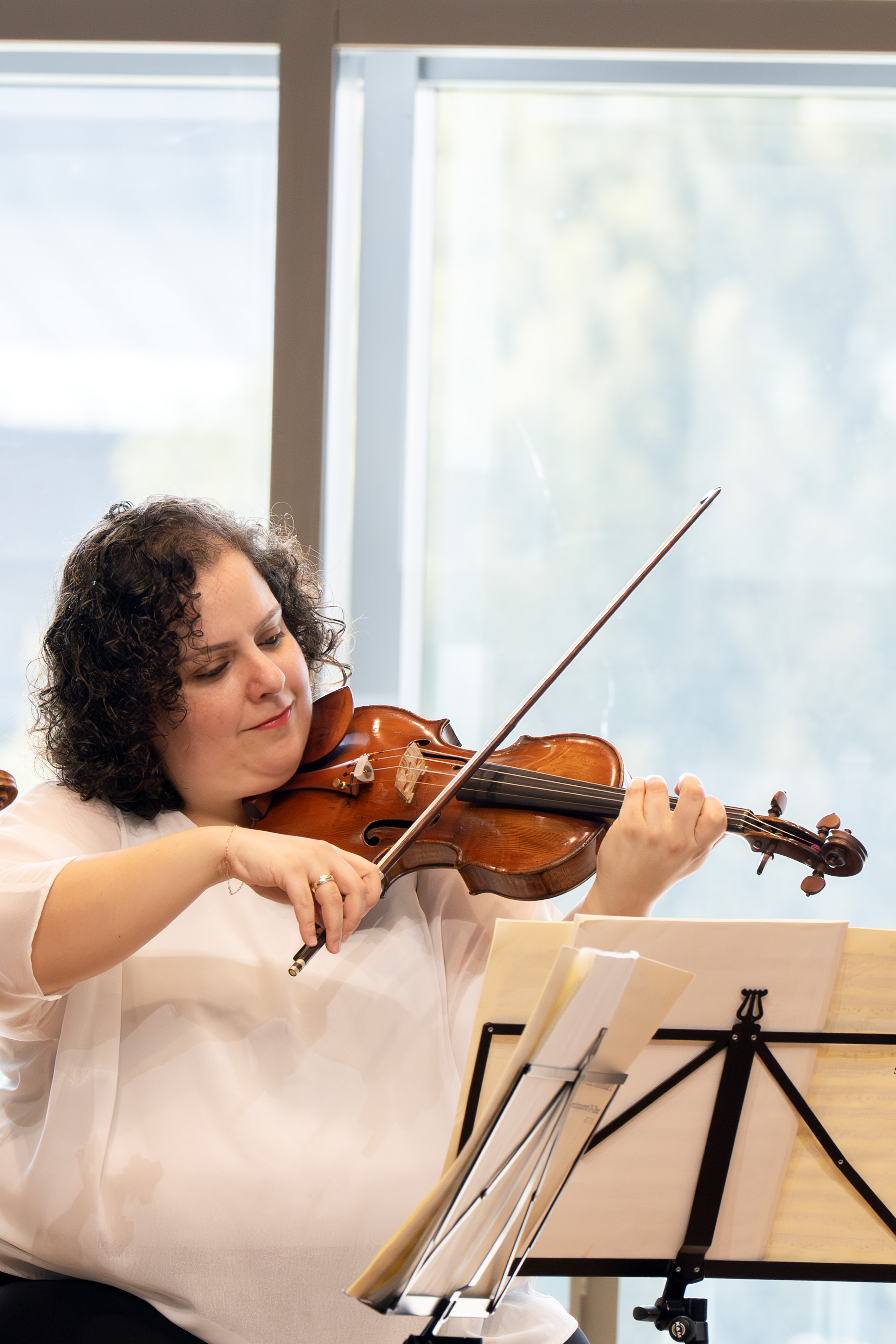 Eine Frau mit lockigem Haar und weißer Bluse spielt Geige und liest Noten auf einem Ständer vor ihr, während große Fenster im Hintergrund natürliches Licht hereinlassen.