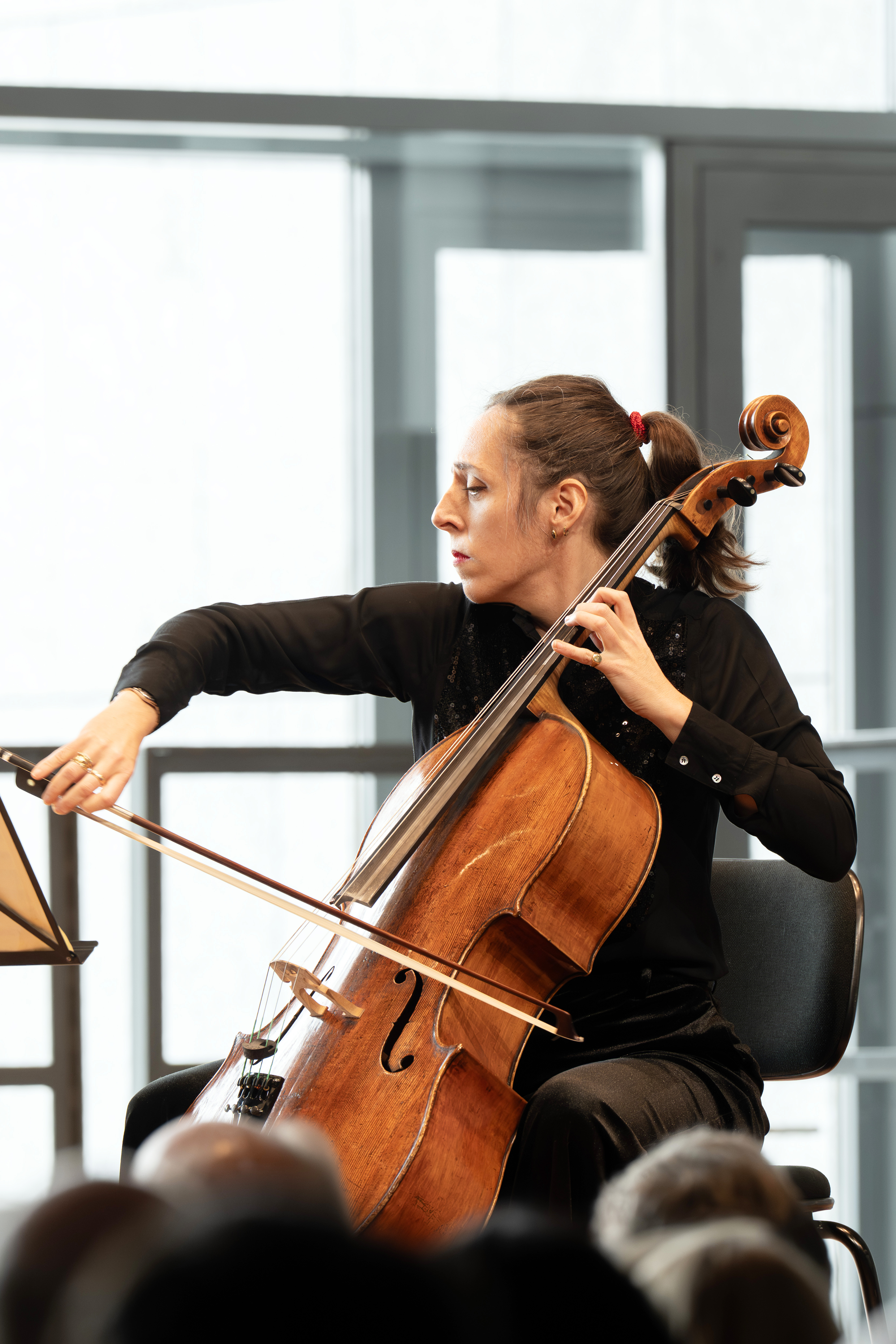 Eine Frau mit Pferdeschwanz spielt konzentriert auf der Bühne Cello und trägt ein schwarzes Outfit. Sie sitzt mit Notenblättern vor sich, und im Hintergrund sind große Fenster zu sehen.